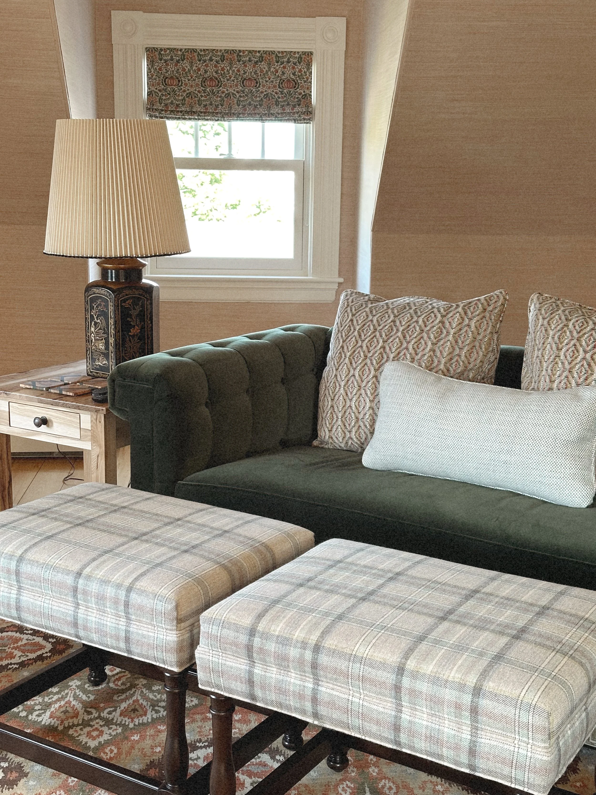 A cozy living room with a green tufted velvet couch, patterned throw pillows, a wooden side table with a lamp, a window with a floral valance, two plaid ottomans, and a patterned rug.