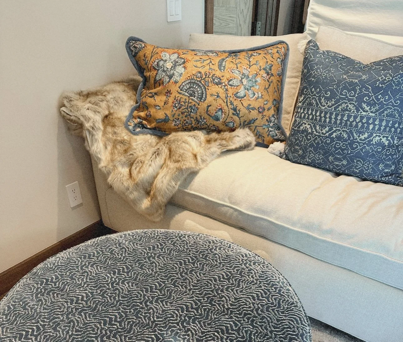 A beige sofa with decorative pillows, a faux fur throw, and a sleeping cat tucked between the pillows.