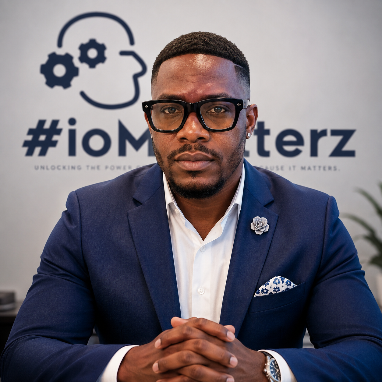A man in a navy suit, white shirt, and glasses sitting at a table. Behind him is a wall with the '#iOMasterz' logo and tagline. The man has a serious expression with his hands clasped.