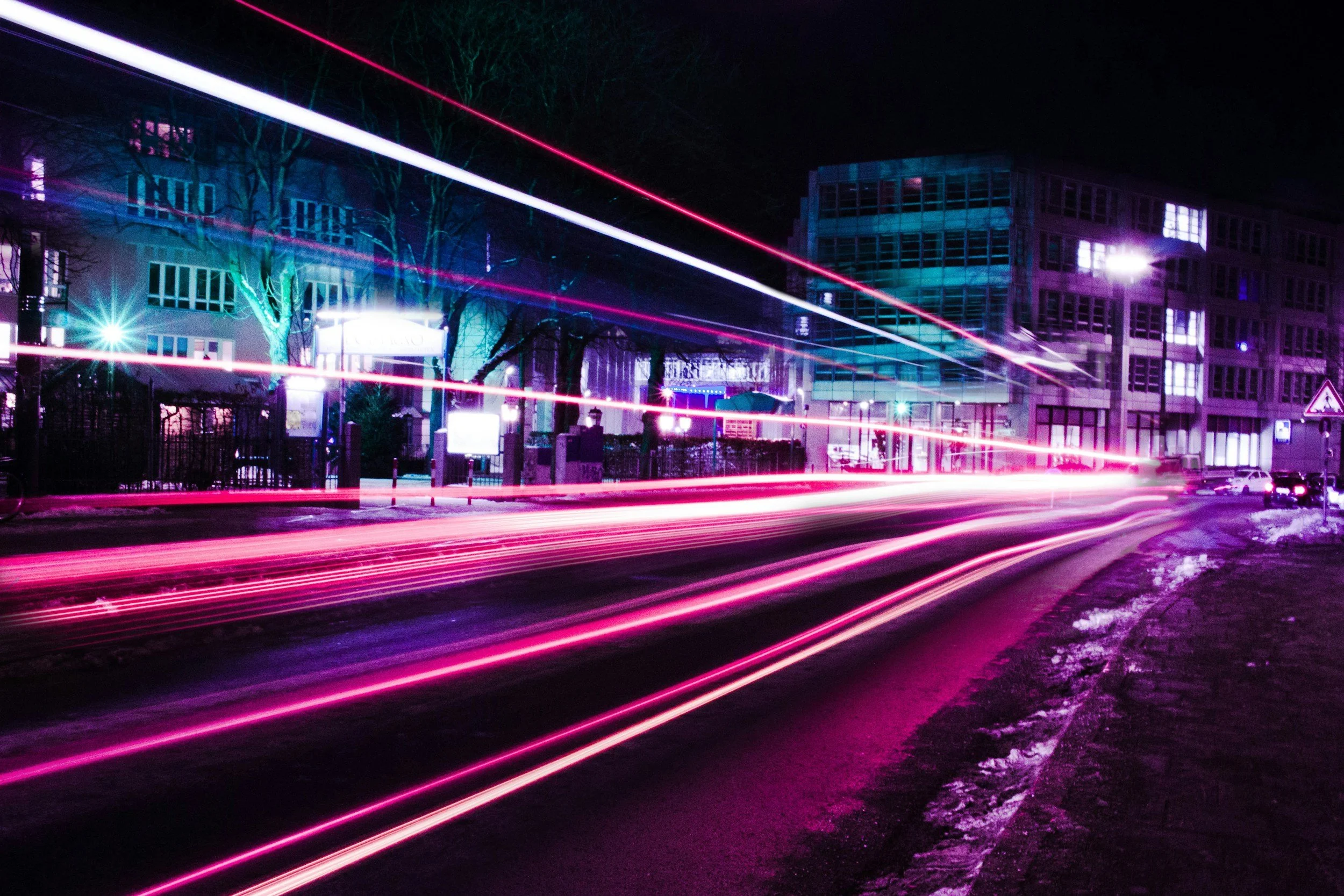 Nighttime city street with light trails from moving vehicles, colorful streetlights, modern buildings, and leafless trees, with some snow on the ground.