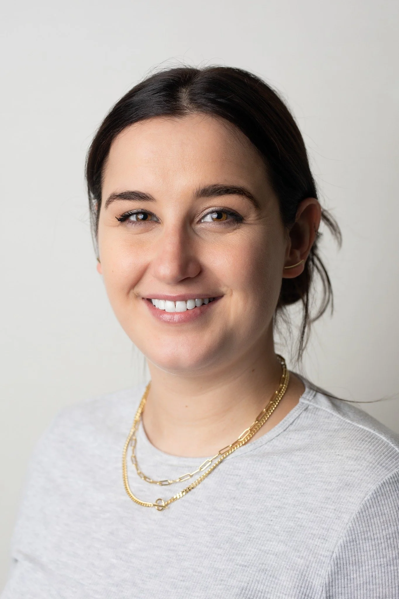 Close-up portrait of a young woman with dark brown hair, smiling, wearing a light gray top and layered gold necklaces, against a plain white background.