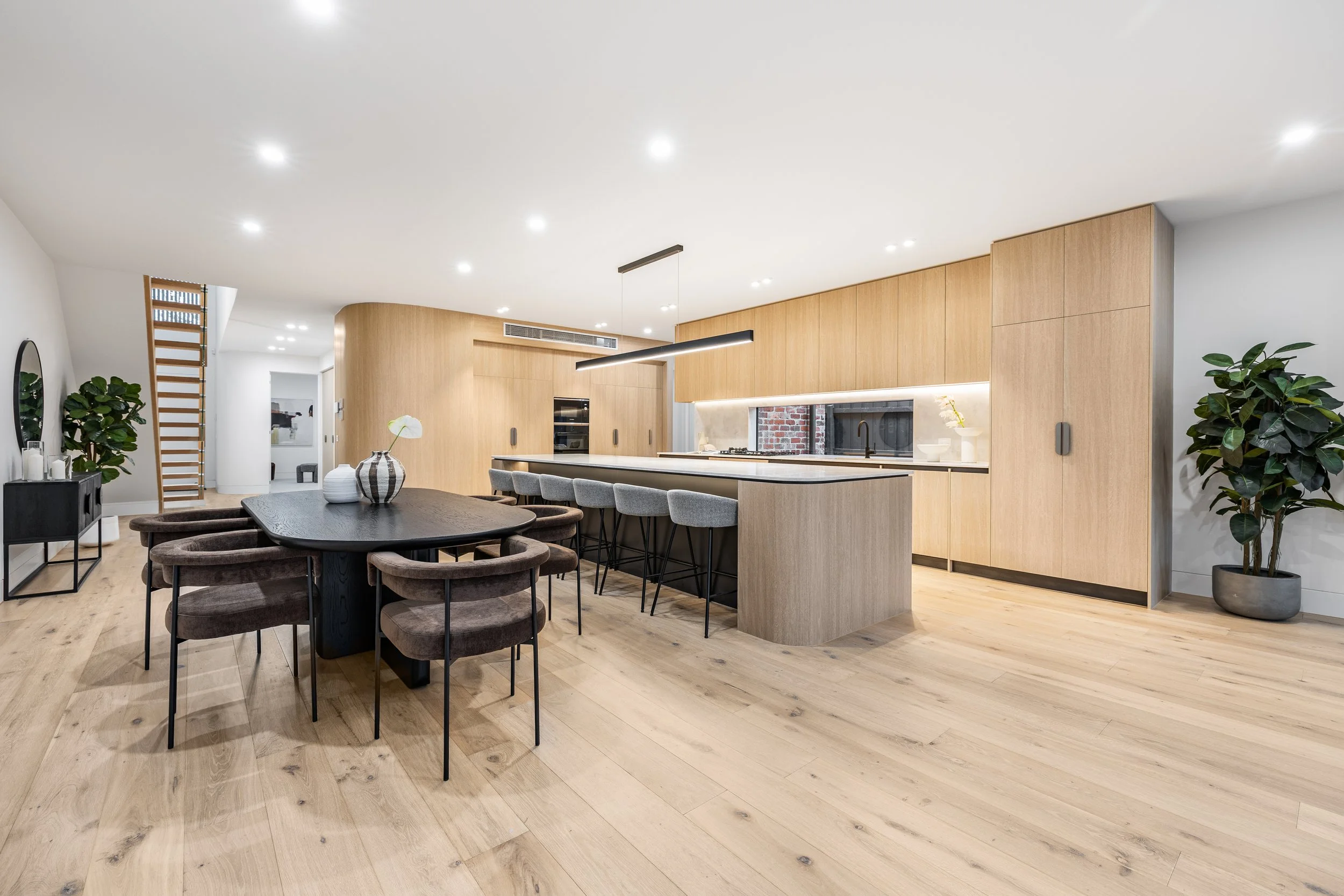 Modern open-concept kitchen and dining area with wood cabinetry, a large black dining table with six chairs, a kitchen island with four bar stools, and potted plants.
