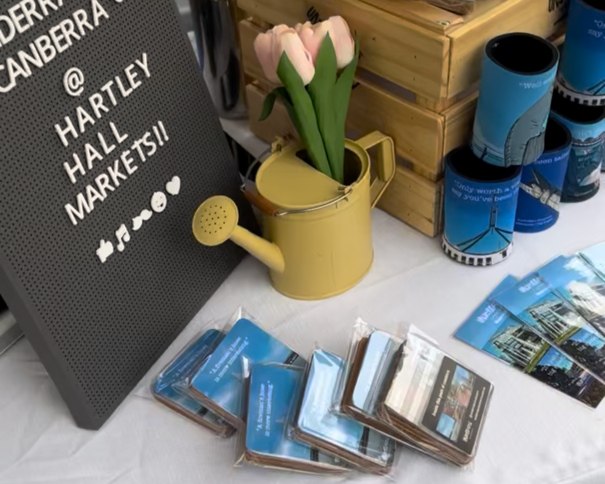 Outdoor market stall under a canopy with a yellow banner reading 'UNDERATED CANBERRA', selling T-shirts, bookmarks, and other items, with a calendar displaying March on the side.