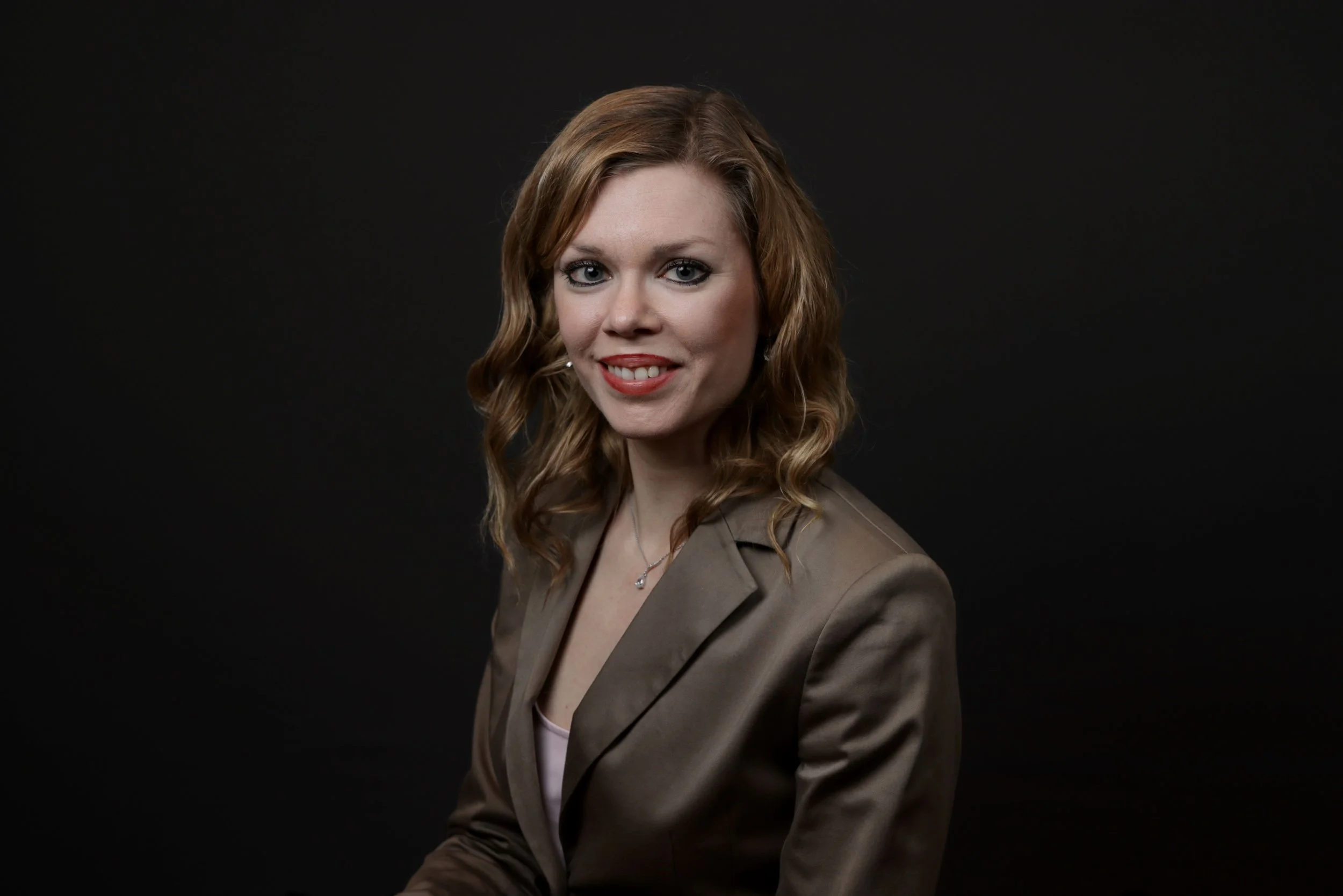 Portrait of a woman with wavy red hair, wearing a brown blazer, smiling against a black background.