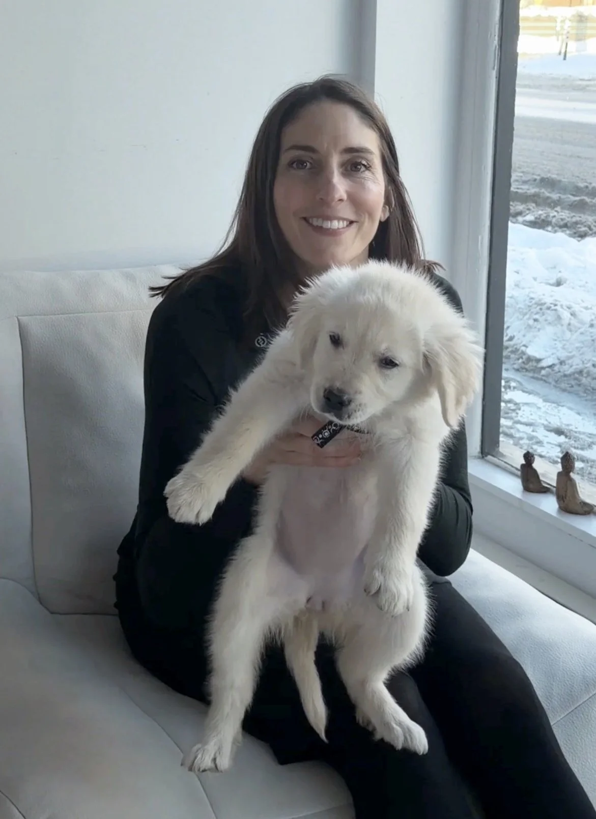 Smiling woman with long dark hair holding a white puppy by a sunny window