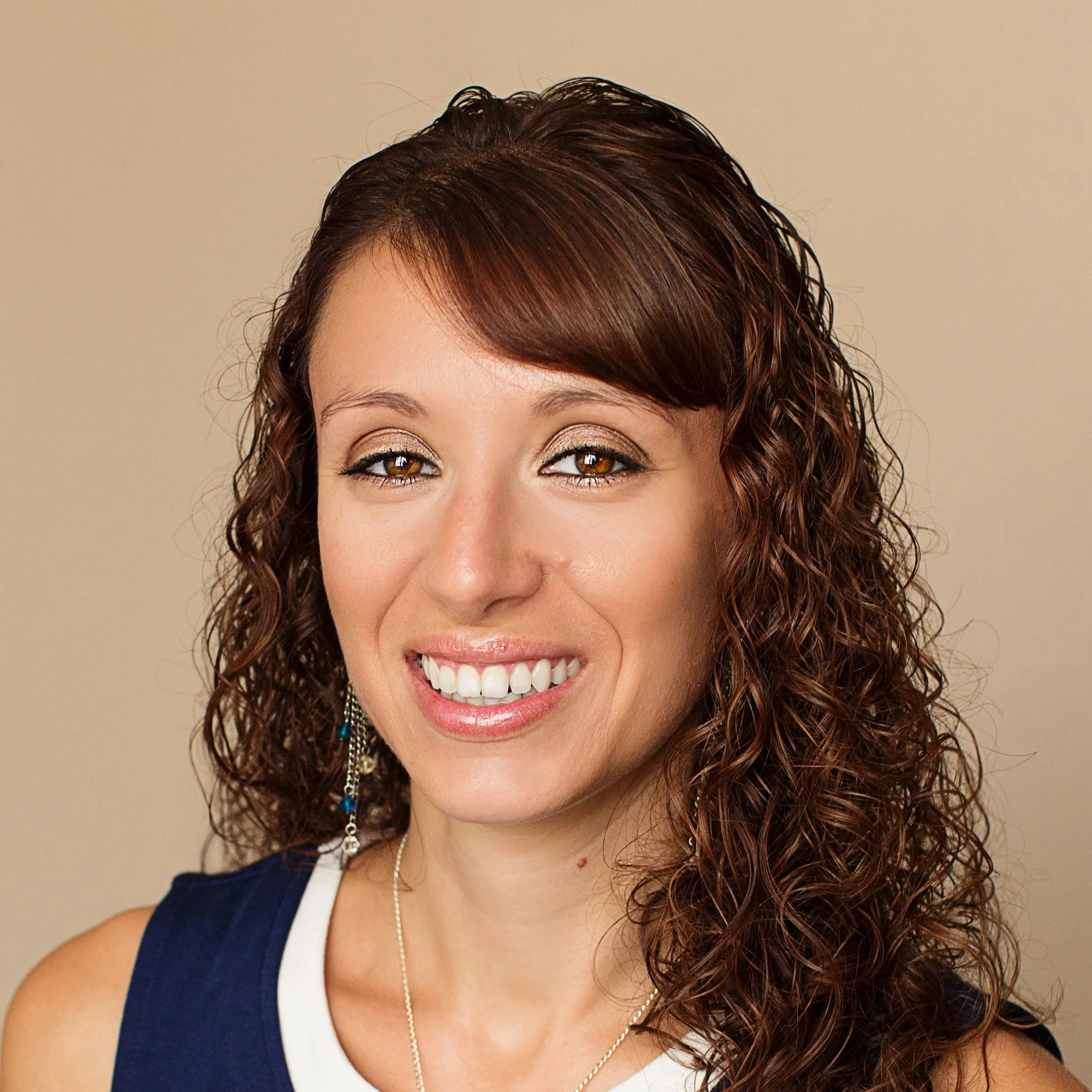 A woman with curly brown hair, wearing a sleeveless navy blue top, smiling, with earrings and a necklace, against a beige background.