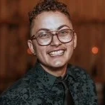 A woman with glasses and curly hair smiling at the camera, wearing a dark patterned shirt.