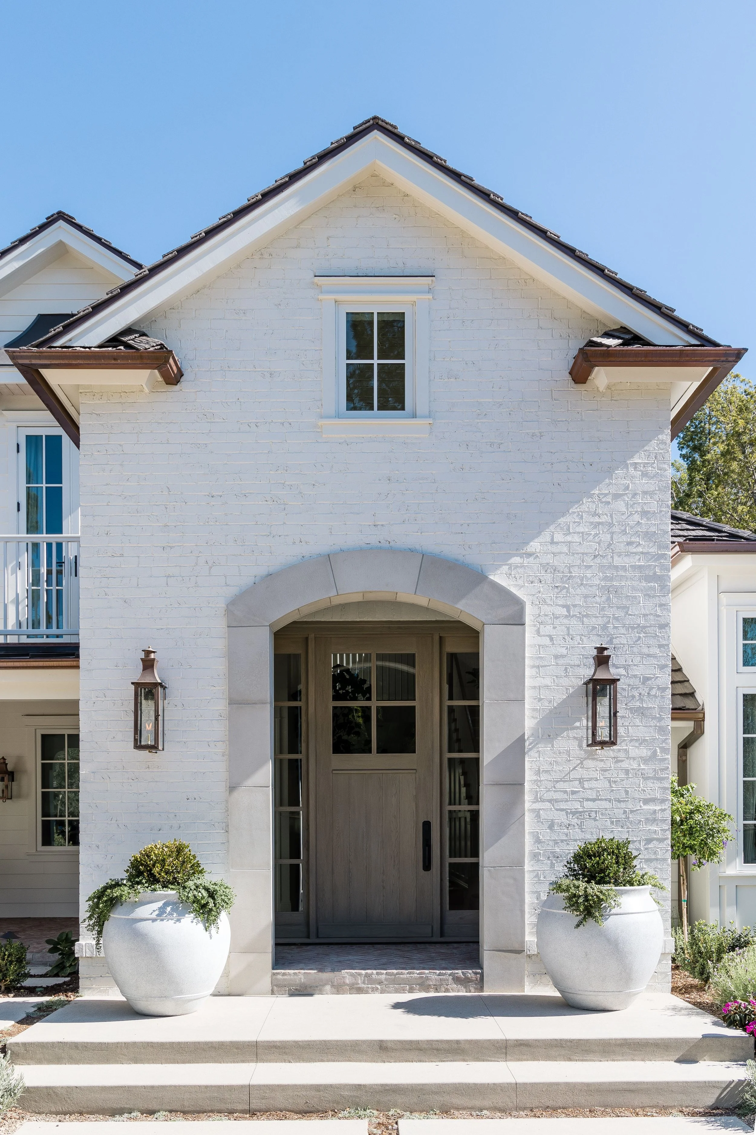 Cedar Front door view of a white brick house with a large arched doorway, two potted plants flanking the entrance, and a window above. The house has a gabled roof with brown shingles and decorative gutters. The sky is clear and blue.