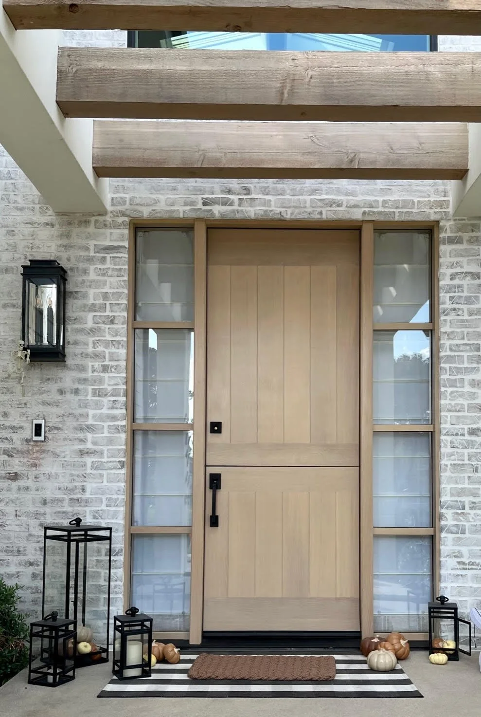 Rift White Oak front door of a house with a natural wood finish, flanked by two large glass side panels, situated on a porch with a striped black and white outdoor rug, decorative lanterns.
