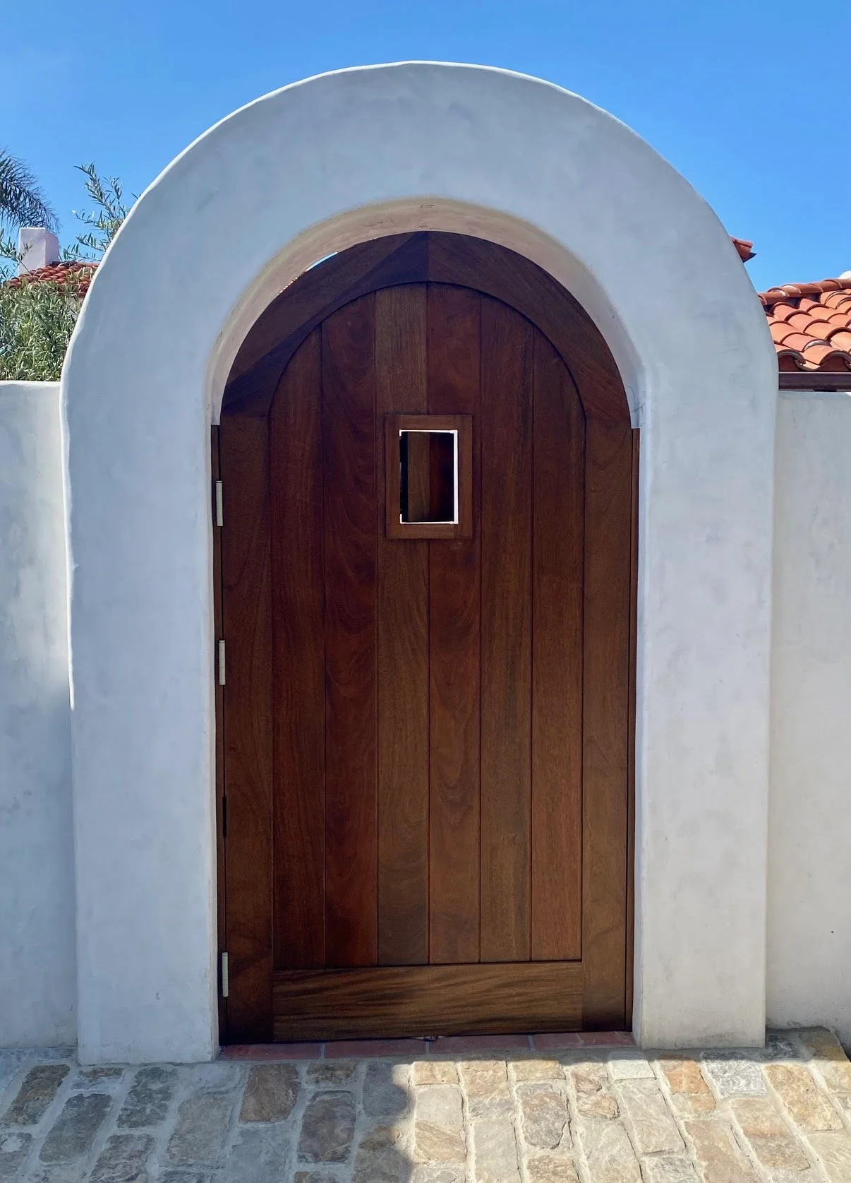 Spanish style, wooden arched door with a small speakeasy window, set in a white stucco wall with a cobblestone ground and red tile roof in background.