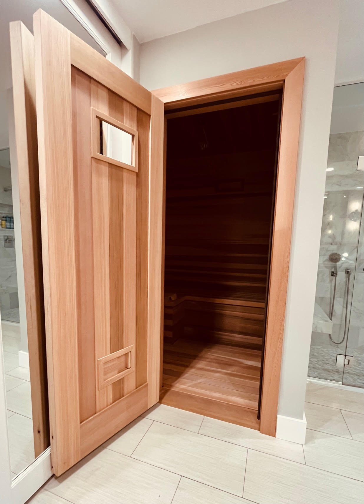 A wooden sauna with an open door showing horizontal wood panels inside, situated next to a modern bathroom with a glass shower.
