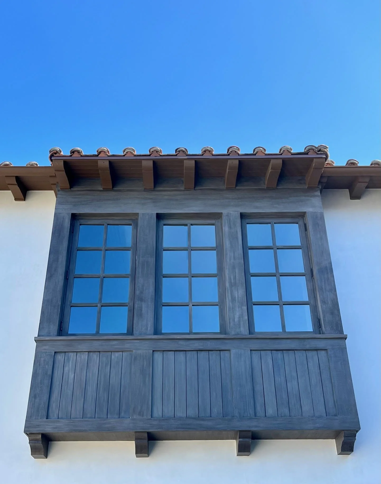 Close-up of a building's exterior corner with large window panels, wooden beams, tiled roof, and white wall, under a clear blue sky.
