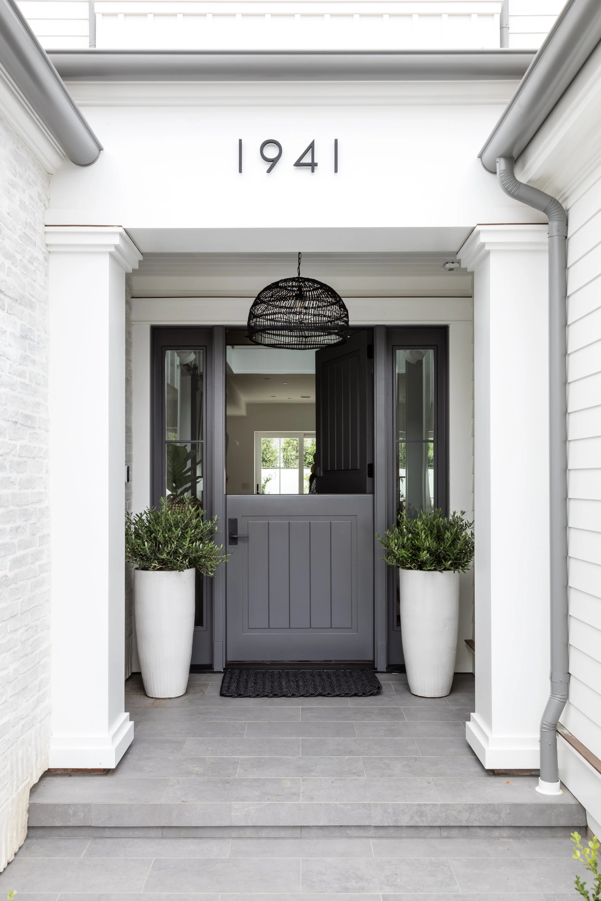 Front door with house number above, two large white planters with greenery on either side, black door with glass panels, black woven pendant light, grey tiled steps, white sides, and a partial view of neighboring house siding.
