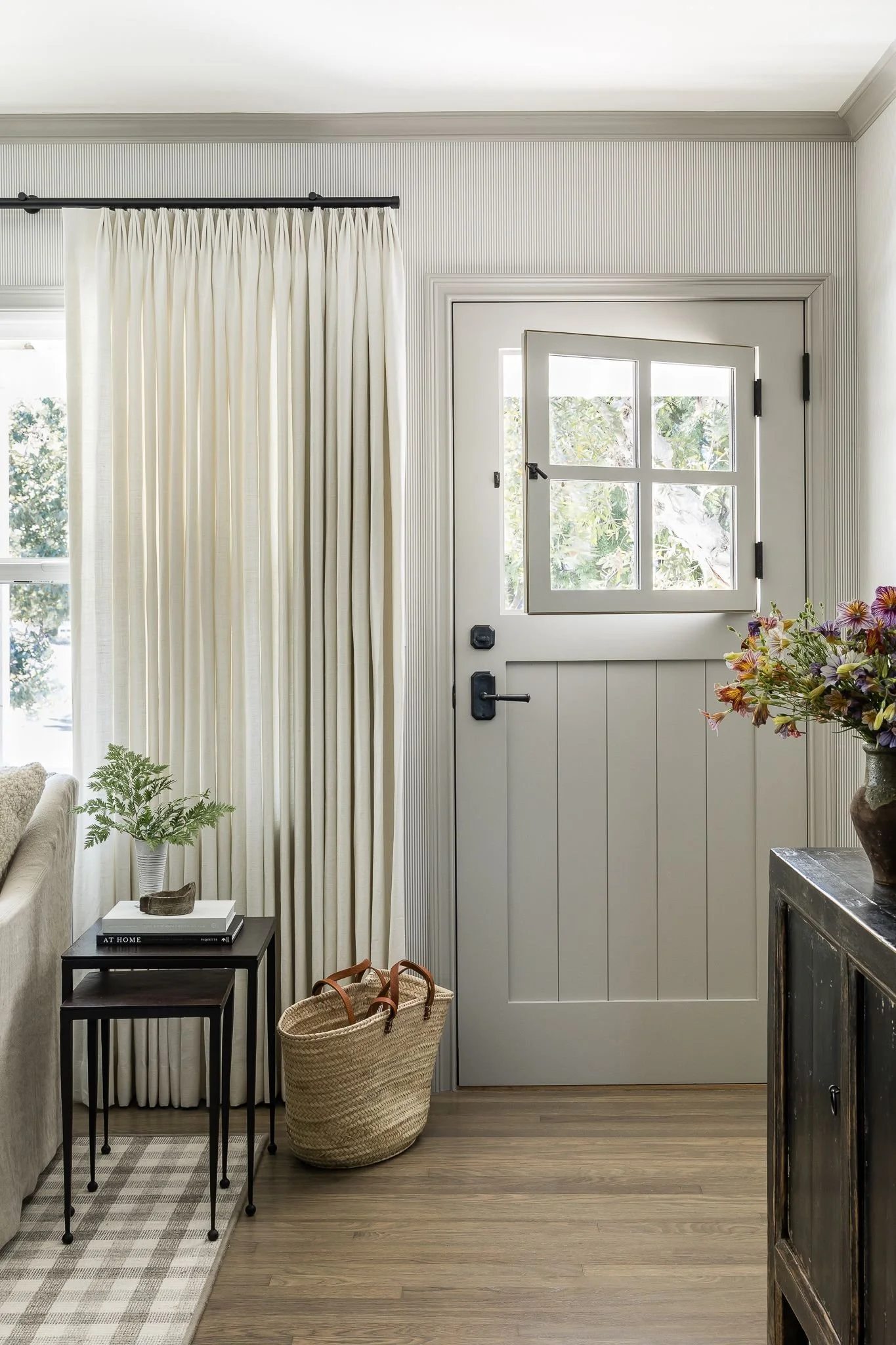 Entryway with white door and window, beige curtains, small black side table with plants and books, woven basket on floor, flowers in a vase, and a light-colored rug.