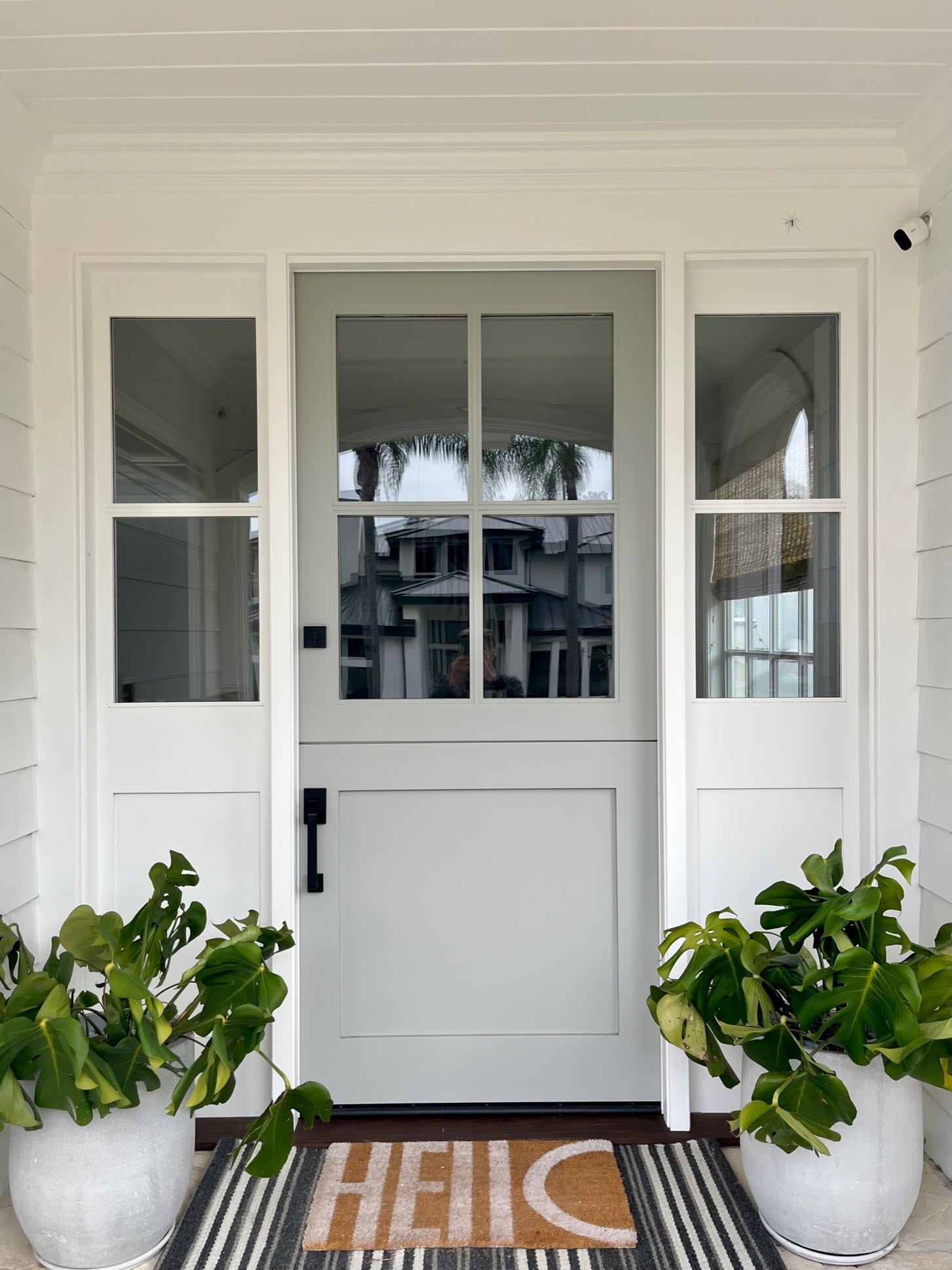 White front door with glass panels, black handle, and deadbolt, flanked by two matching white planters with green plants, and a striped doormat with the word 'HI' on it, outside a house.
