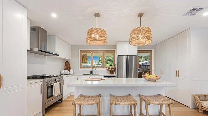 Modern kitchen with white cabinetry, stainless steel appliances, two woven pendant lights, a white island with three wicker stools, and a window overlooking a green outdoor area.