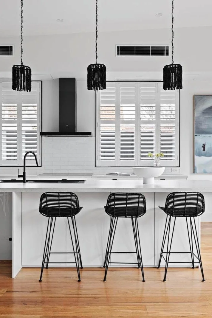 Modern kitchen with white island counter, black bar stools, black pendant lights, white window shutters, a bowl with flowers, and a piece of wall art.