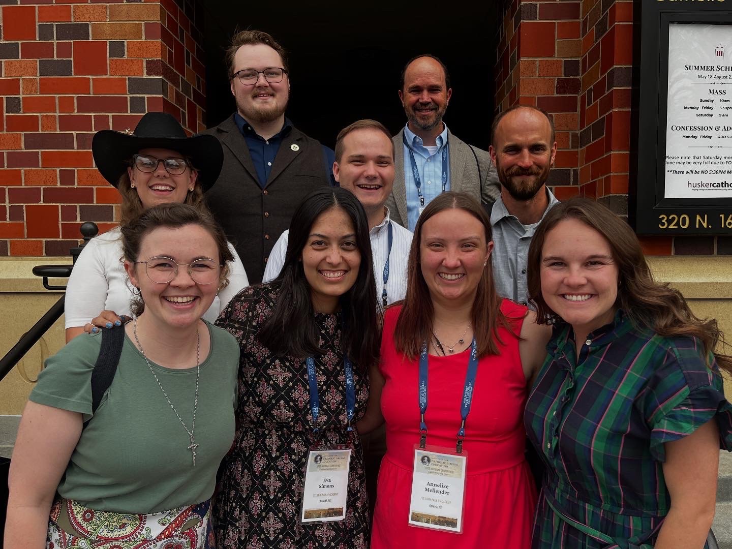Group of ten people smiling in front of a brick building, some wearing conference name tags, with a sign on the right side of the image.