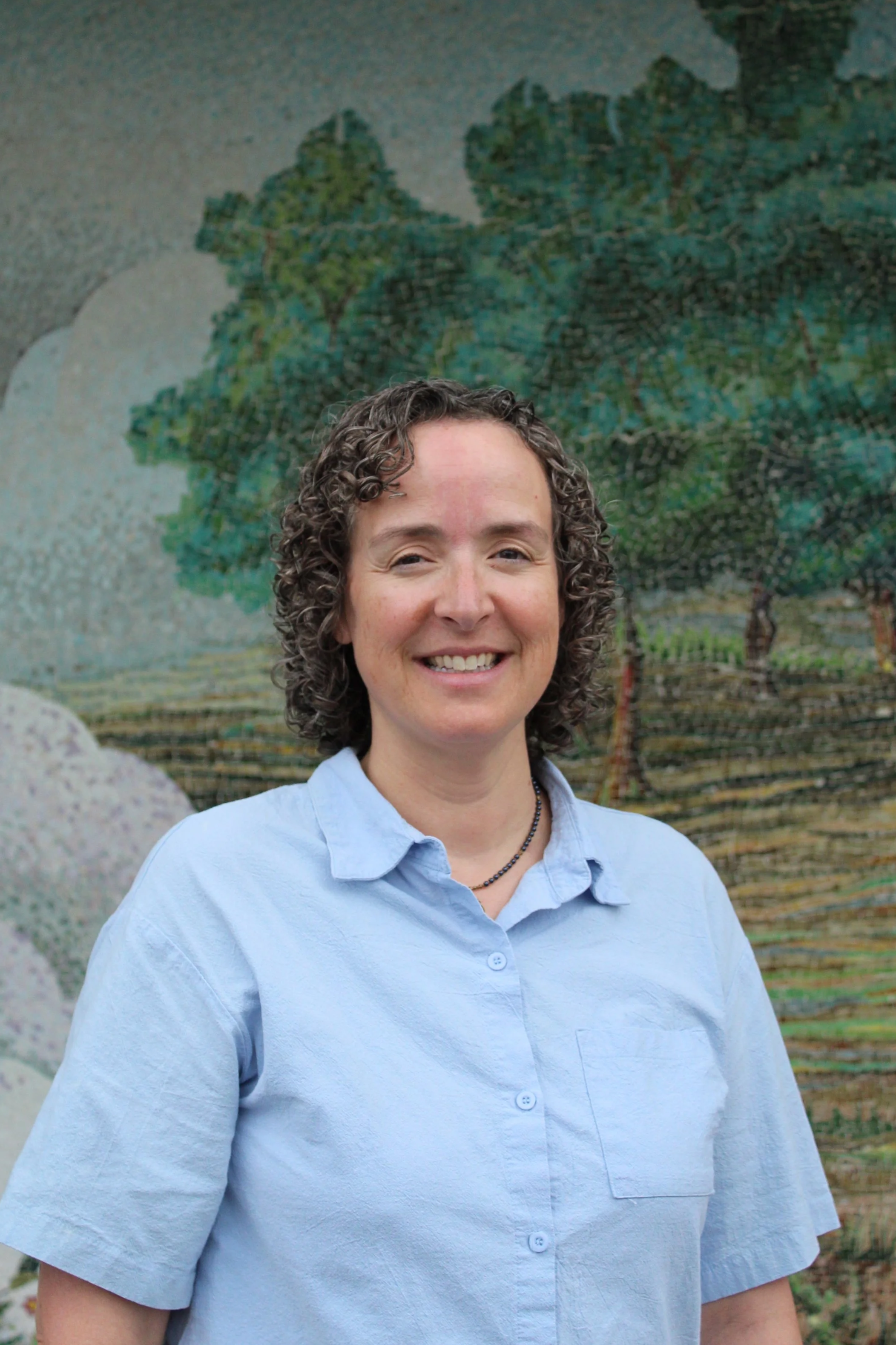 A woman with short, curly gray hair smiling, wearing a dark blue cardigan over a white blouse with a blue leaf pattern, and layered necklaces, standing in front of a colorful mosaic artwork depicting trees and nature.