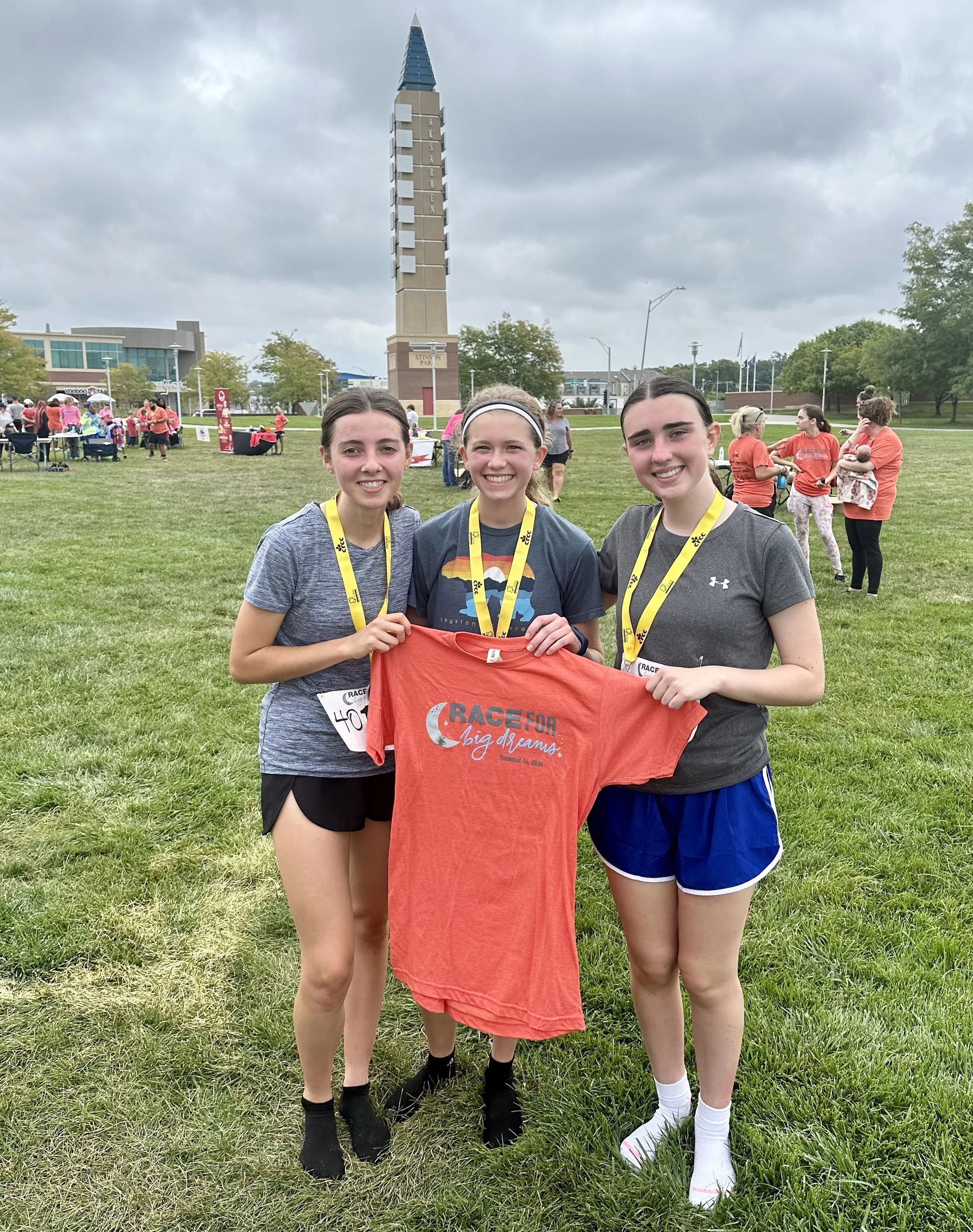 Three young women standing on grass at a race event, holding a red t-shirt with the text 'Race for Big Dreams' in front of a tall clock tower and other participants in the background.