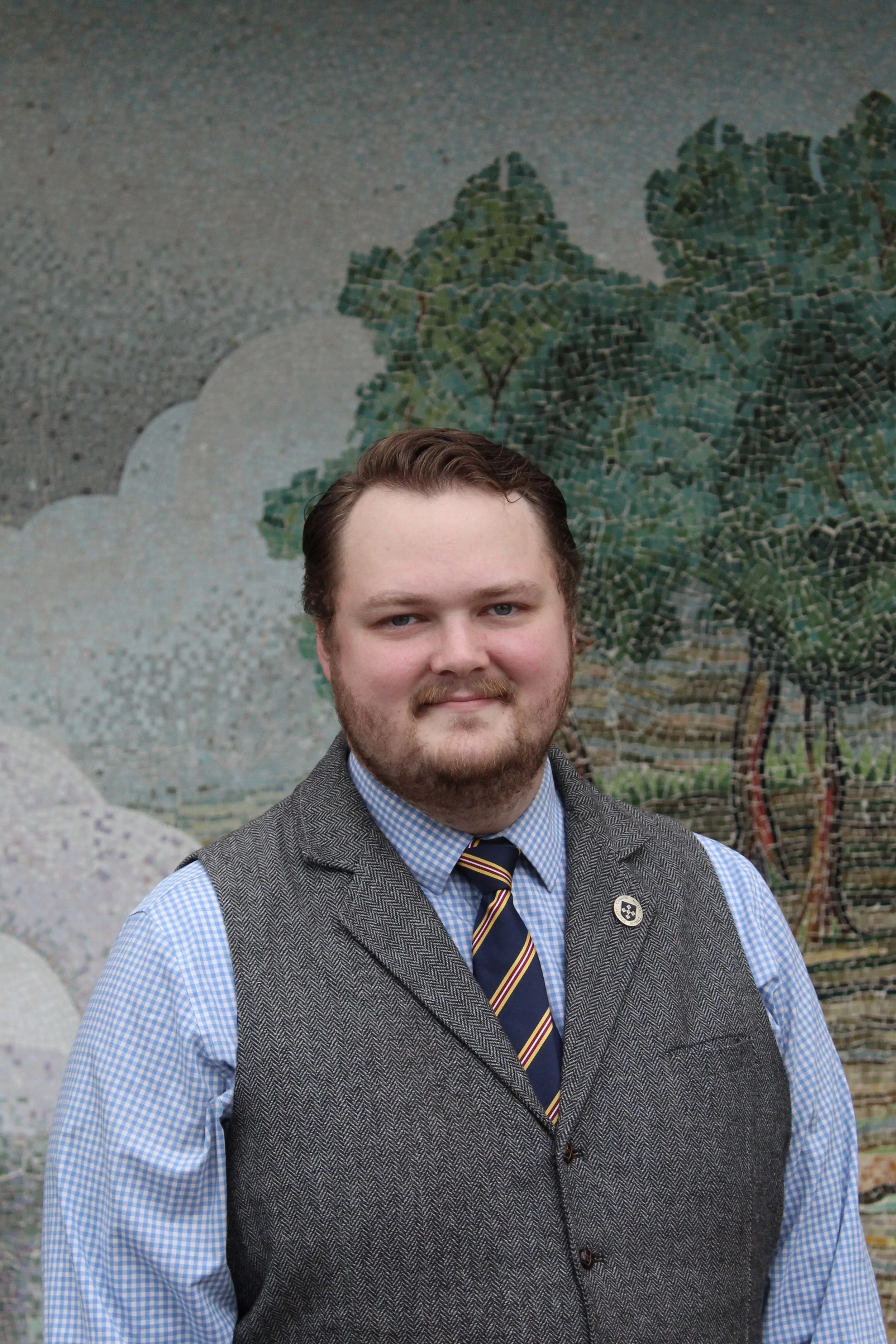 Young man in glasses and a suit standing in front of a colorful mural of trees.
