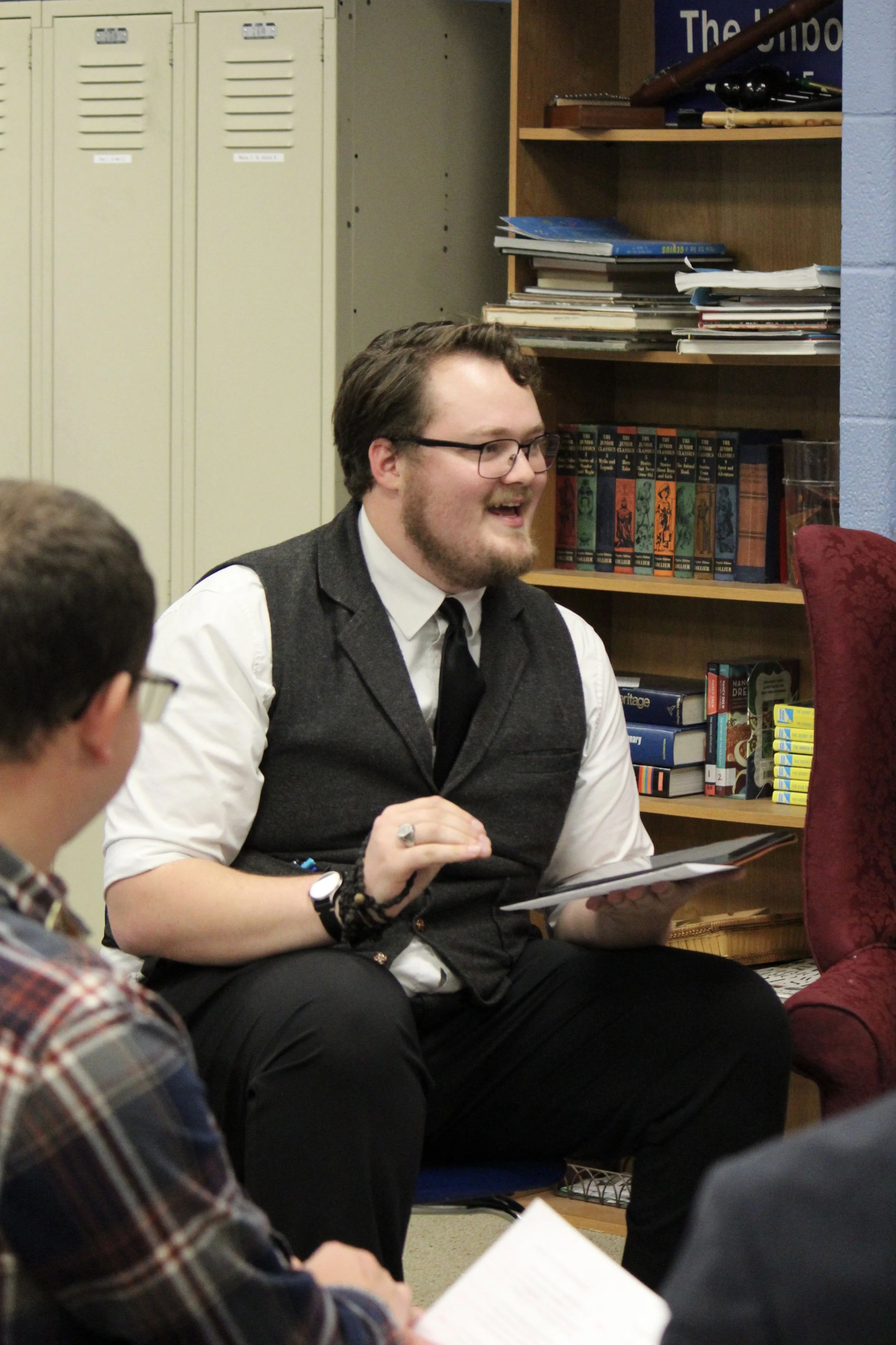 A young man with glasses, a beard, and curly hair, wearing a white shirt, black tie, and a gray vest, is seated and smiling while holding a tablet in a room with bookshelves and a red upholstered chair nearby.
