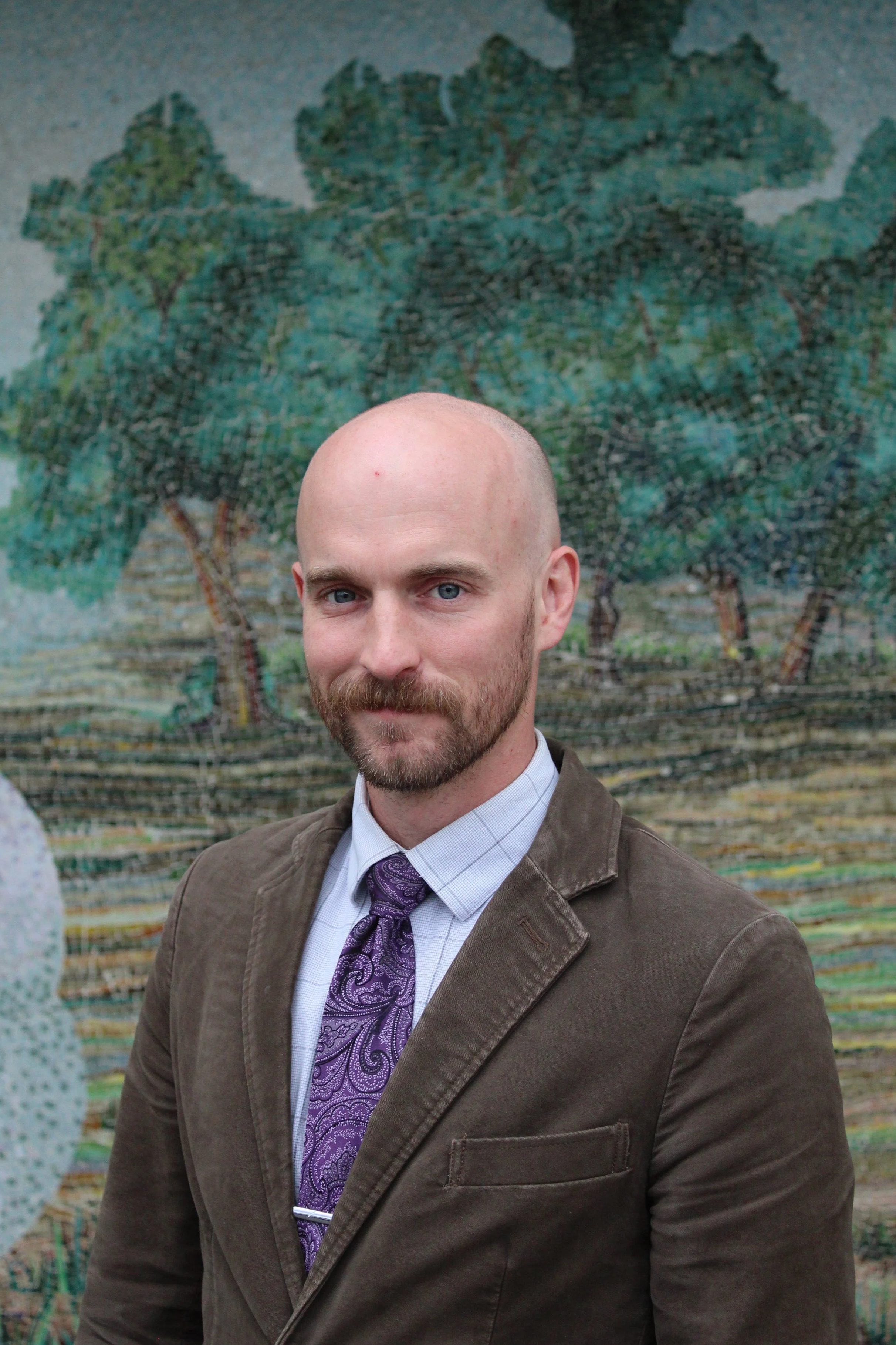 A man in a dark suit, light blue shirt, and black tie standing outdoors in front of a colorful mural with trees painted on it.