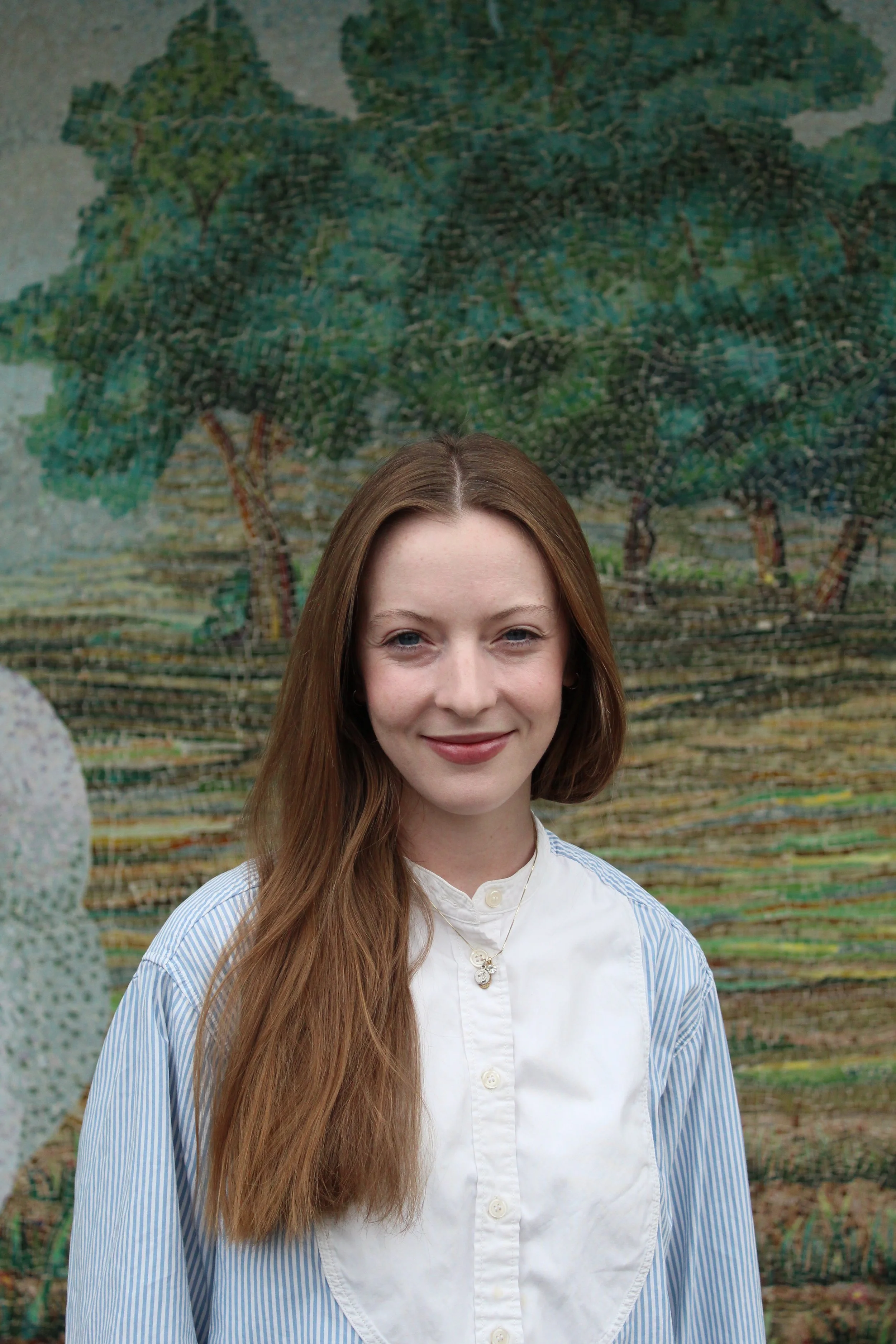 A young woman with long brown hair, wearing a sleeveless black turtleneck top and gold hoop earrings, standing outdoors against a large, green, leafy tree mural background.