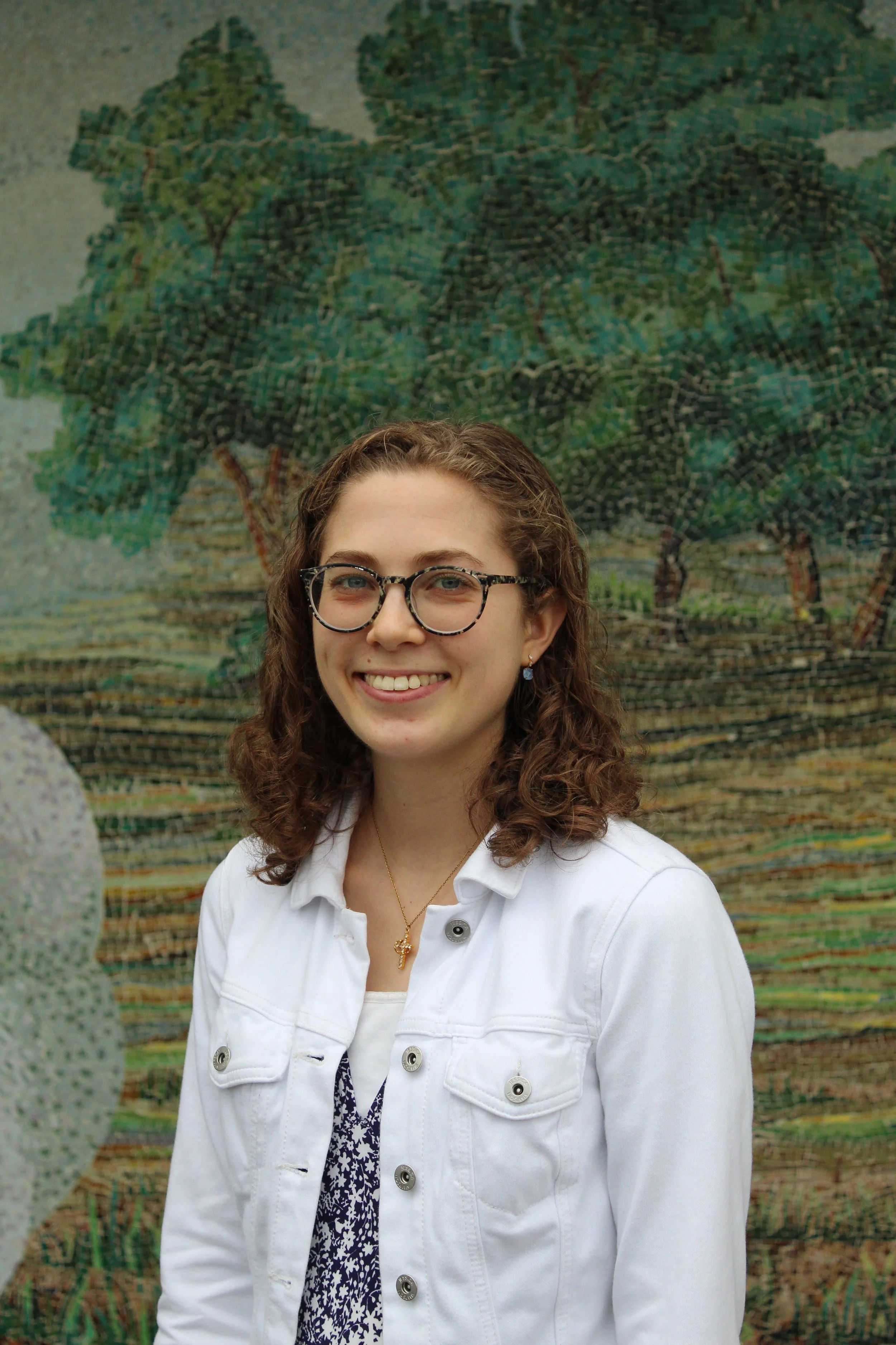A young woman with curly brown hair, glasses, and a white jacket is smiling at the camera, standing in front of a colorful landscape painting of trees and a scenic outdoor scene.