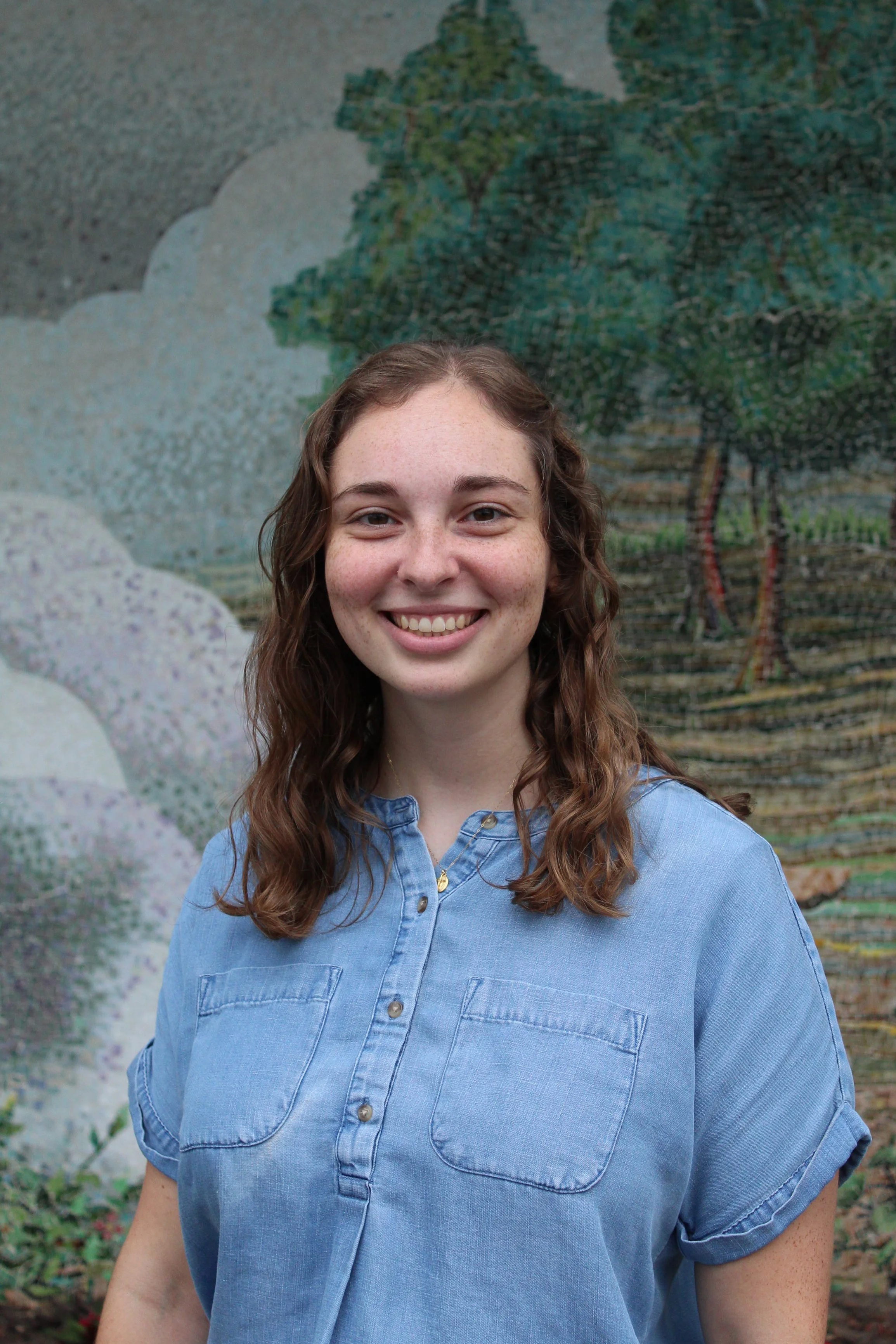 Young woman with long wavy brown hair, smiling, wearing a blue button-up shirt, standing in front of a colorful outdoor mural of trees.