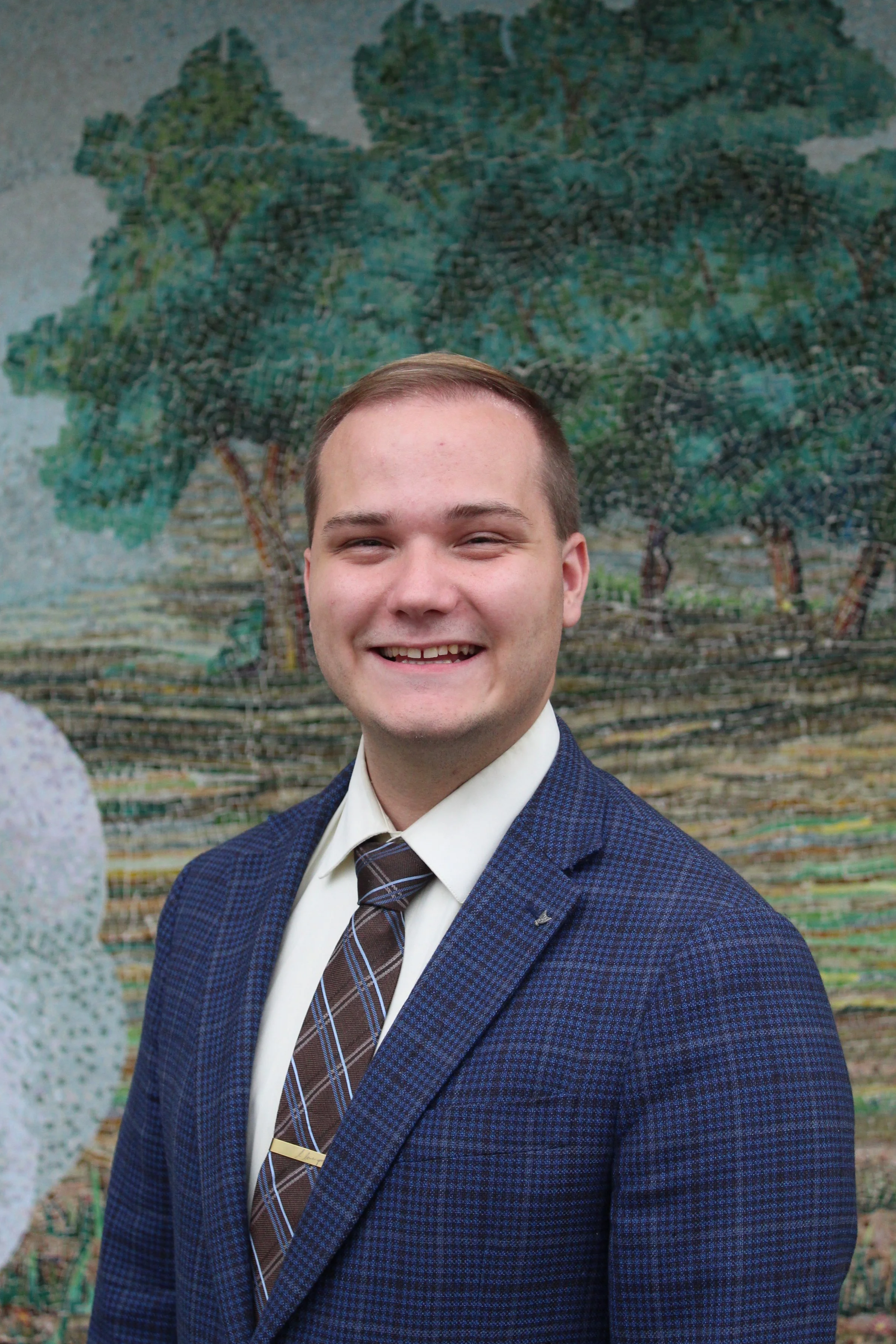 A young man with short brown hair, smiling, wearing a blue checkered blazer, light blue shirt, and striped tie, standing in front of a mural featuring a large tree with green leaves and a textured background.
