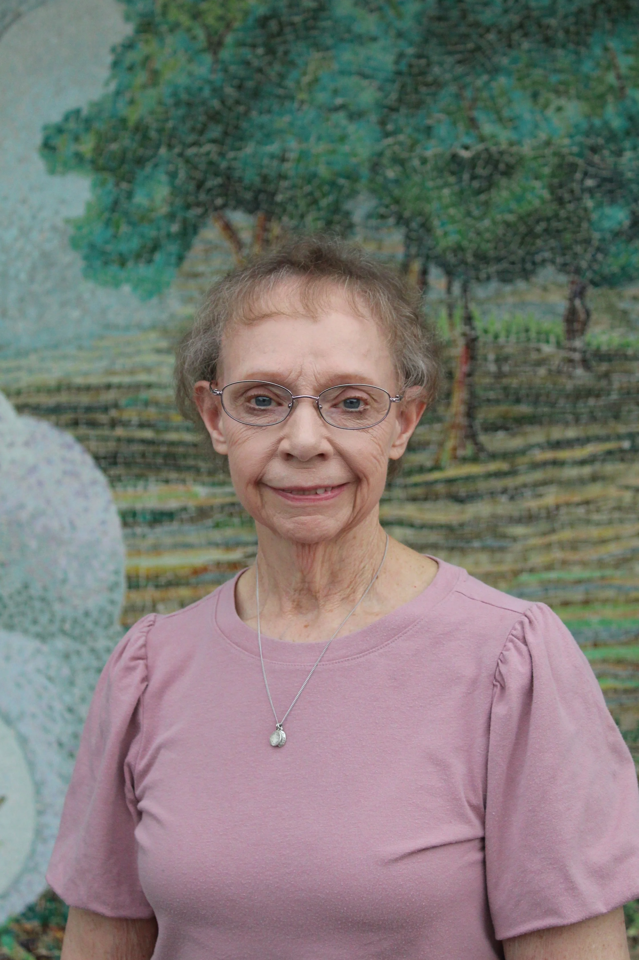 An elderly woman with short hair and glasses, wearing a red top with button details on the sleeve, stands in front of a colorful mosaic wall.