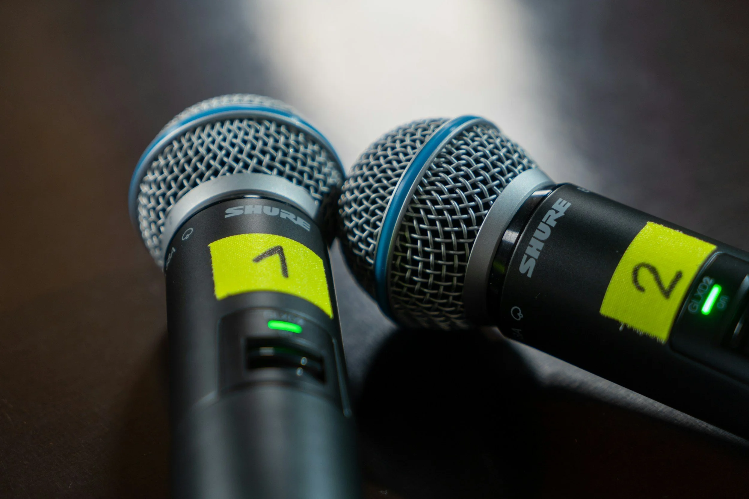 Two Shure wireless handheld microphones with yellow tape labels numbered 1 and 2, placed on a dark surface.