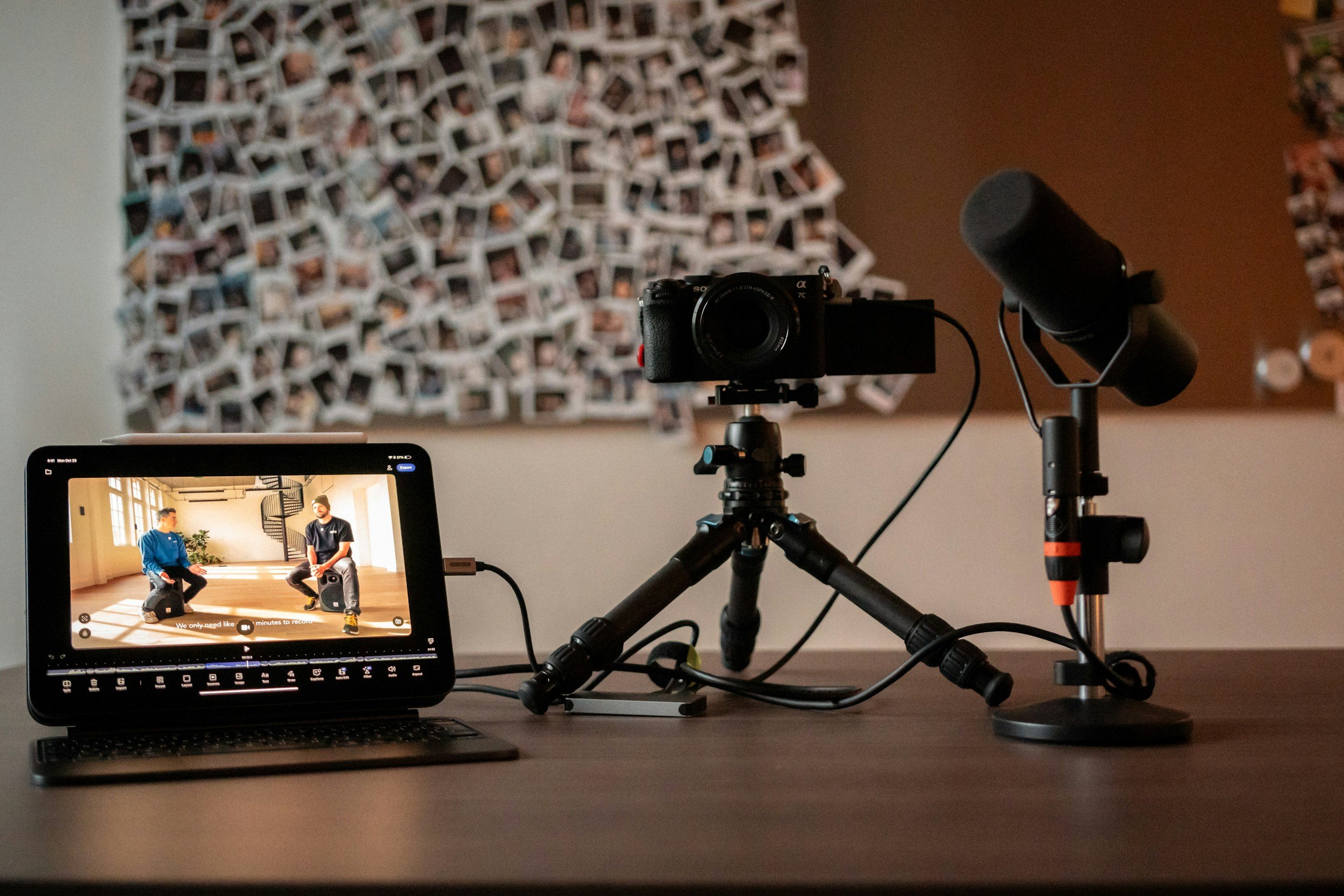 A camera on a tripod is set up facing a monitor showing two men sitting on the floor in a spacious room with large windows and a spiral staircase in the background. The monitor displays a real-time video with a caption