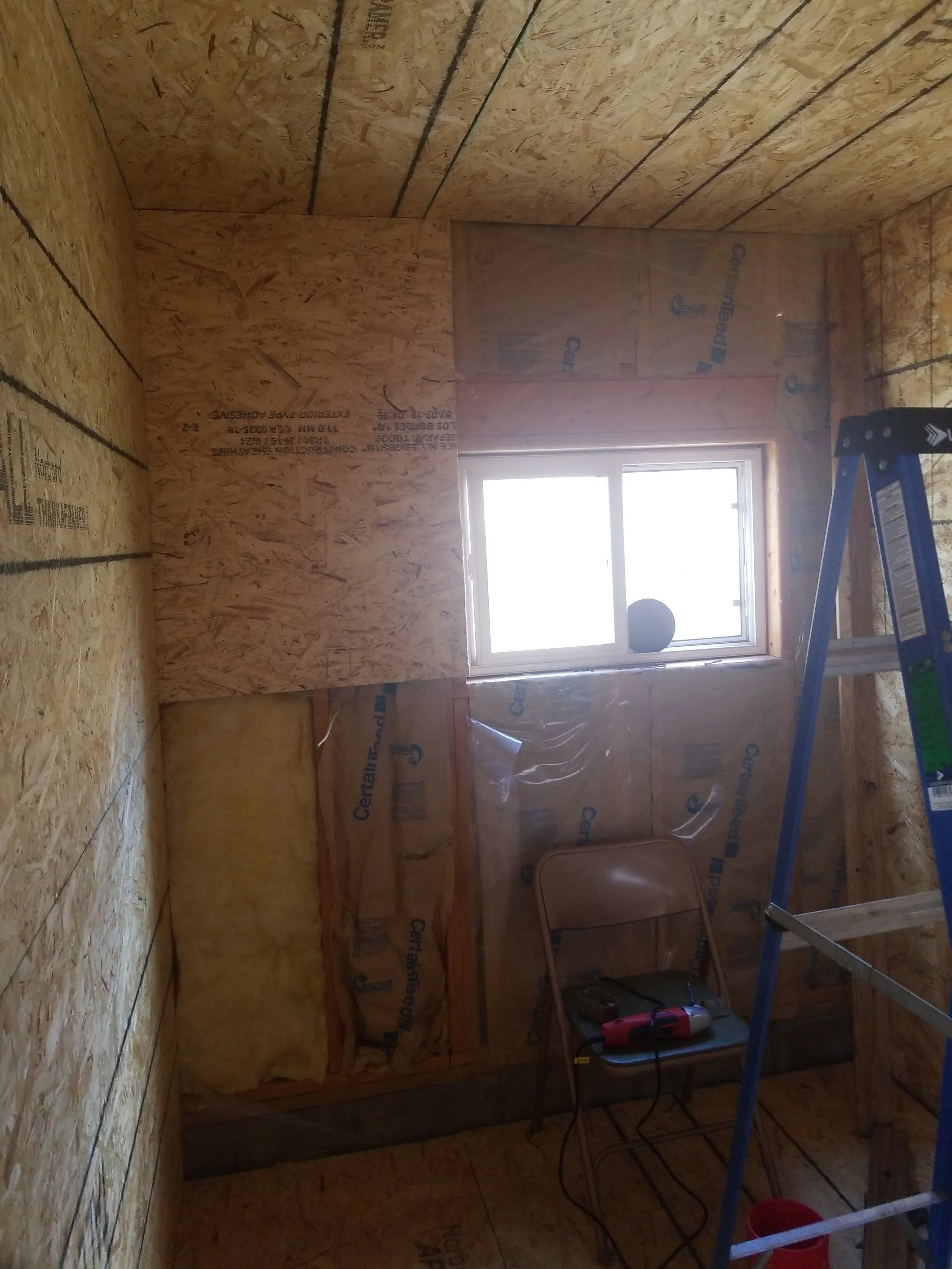 A partially finished room under construction with exposed plywood walls, a small window, a folding chair, a red portable light, and a blue step ladder.