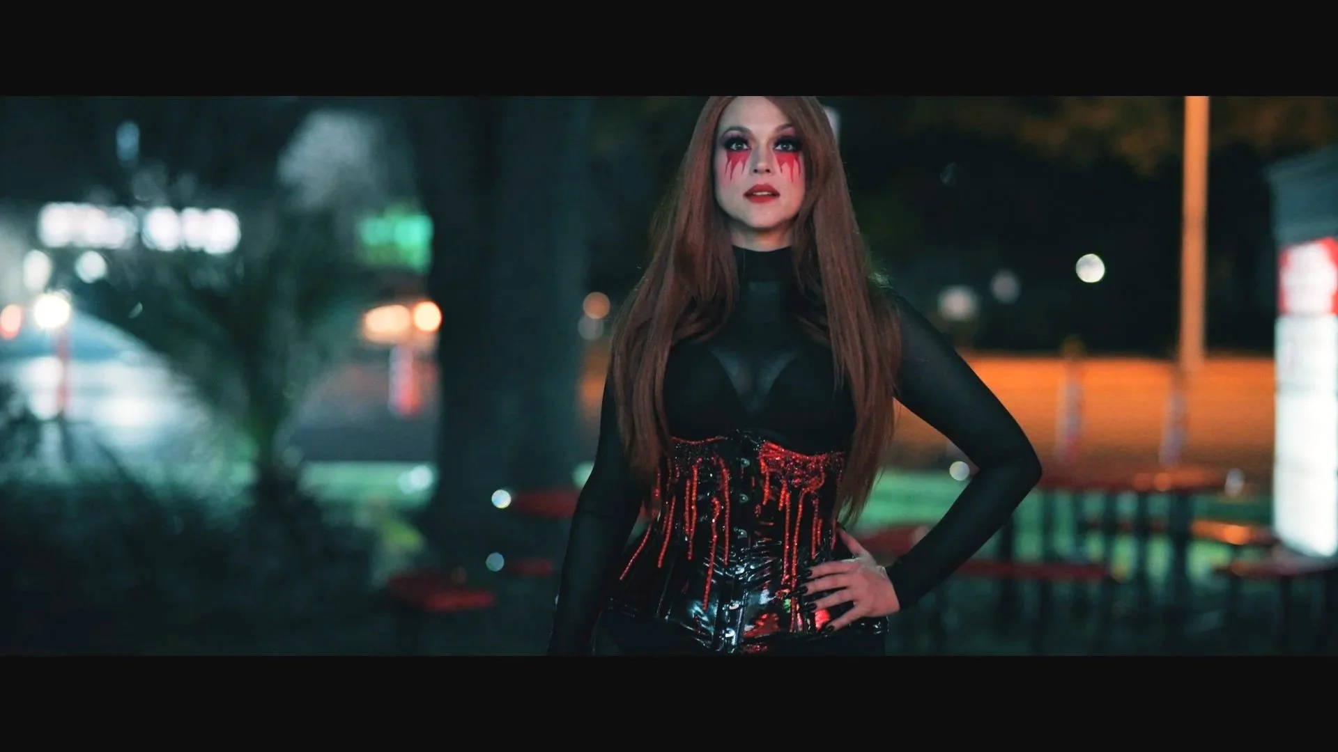 Scarah outdoors at night in a black sheer top and latex skirt with red detailing, hand on her hip