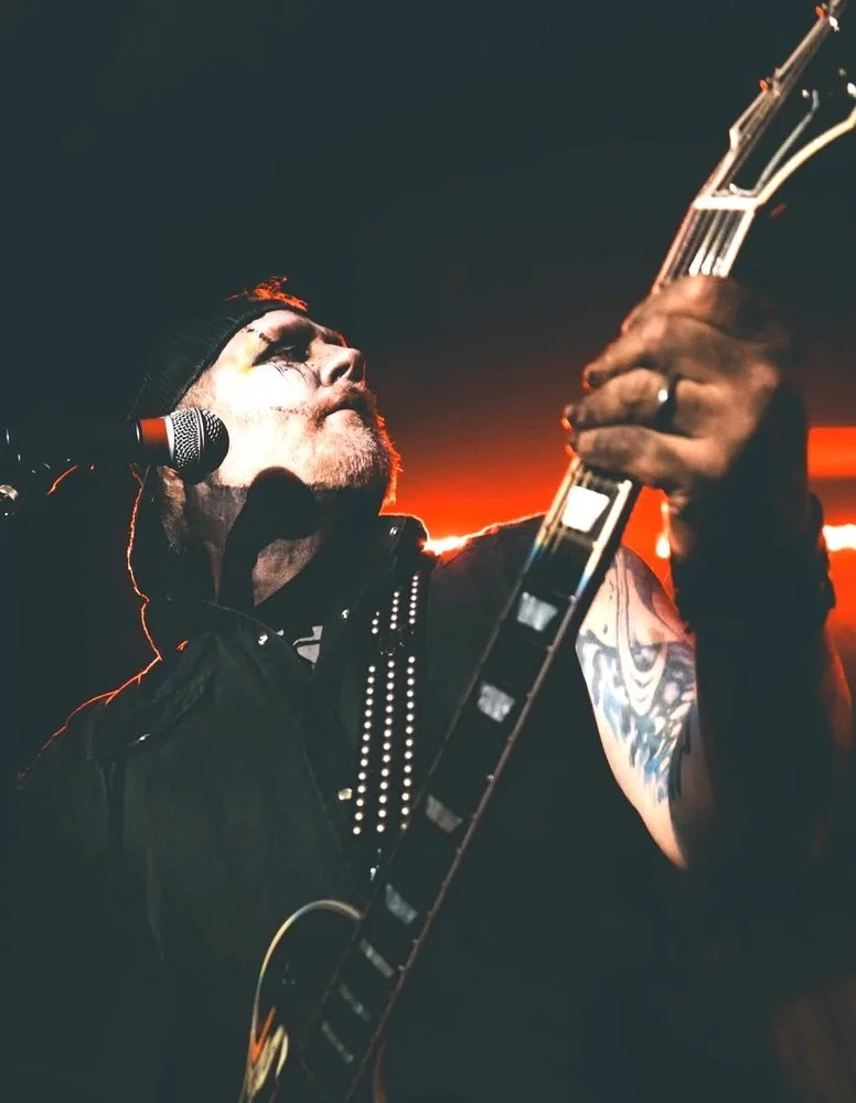 Gorey Corey playing guitar, backward cap and theatrical eye makeup, red backlight