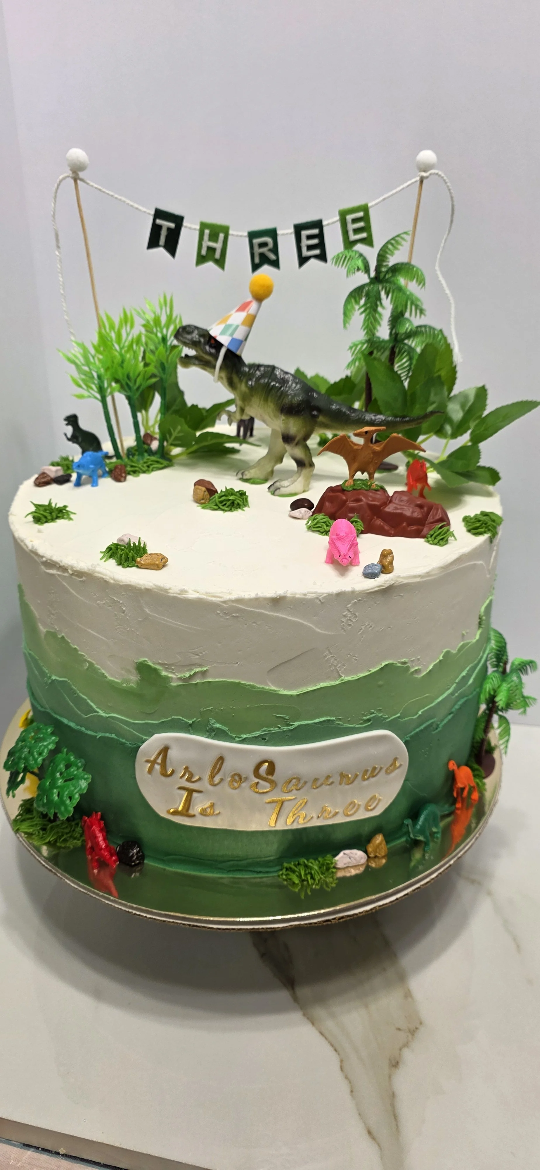 A birthday cake decorated with a dinosaur theme, featuring miniature dinosaurs, plants, rocks, and a banner that reads 'THREE'. The cake has an inscription that says 'Arlo Saunatural Is Throo'.