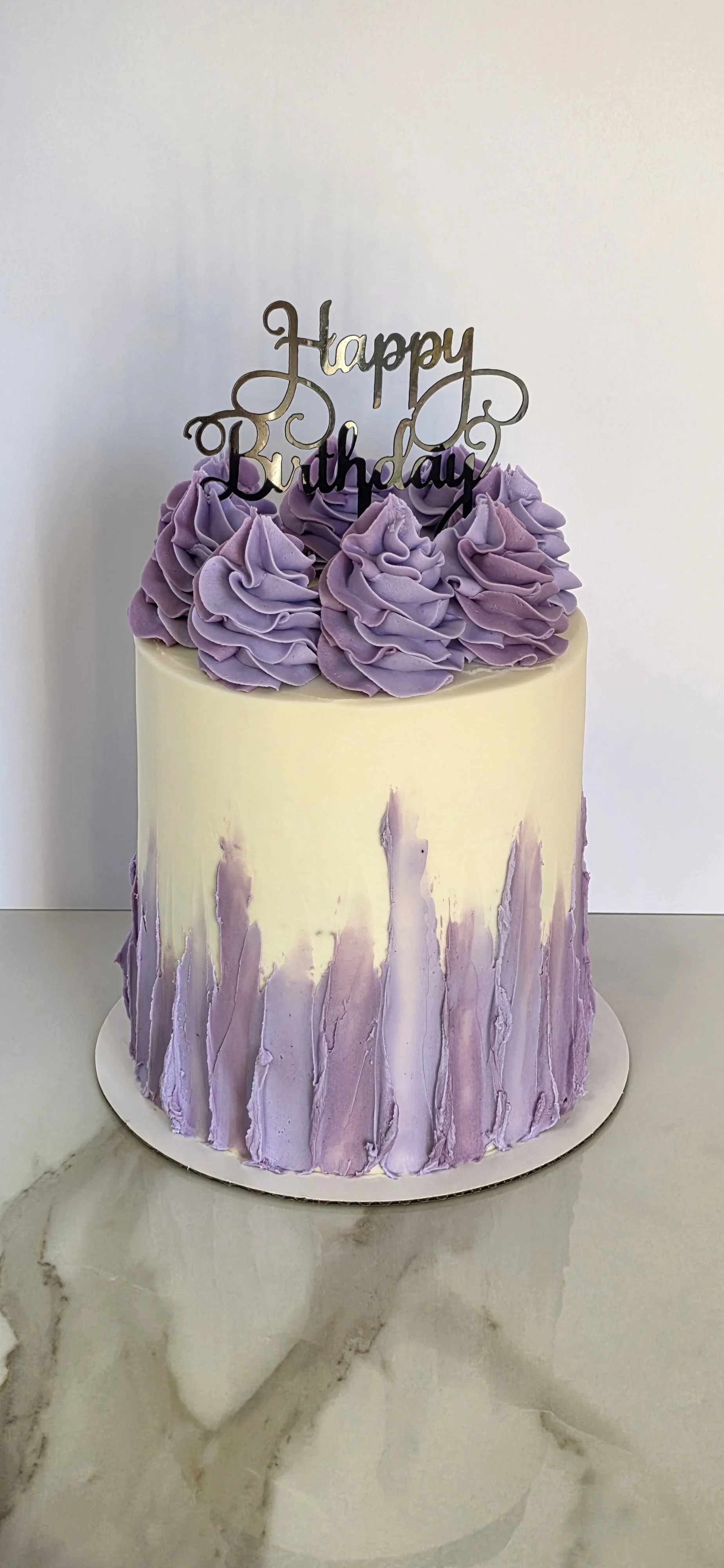 A two-layer birthday cake with purple frosting decorations and a "Happy Birthday" topper, placed on a white marble surface.