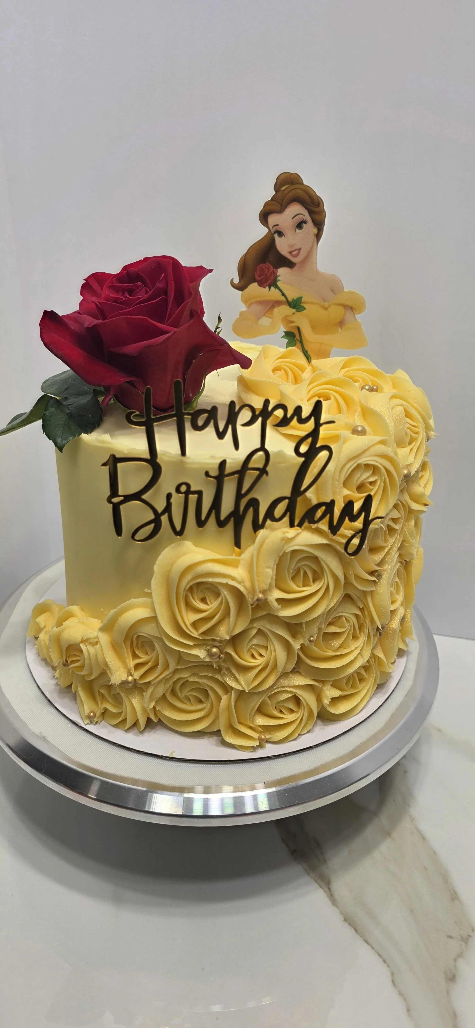 A birthday cake with yellow rose-shaped frosting, a red rose, and a Belle character cutout from Disney's Beauty and the Beast, with a "Happy Birthday" message.