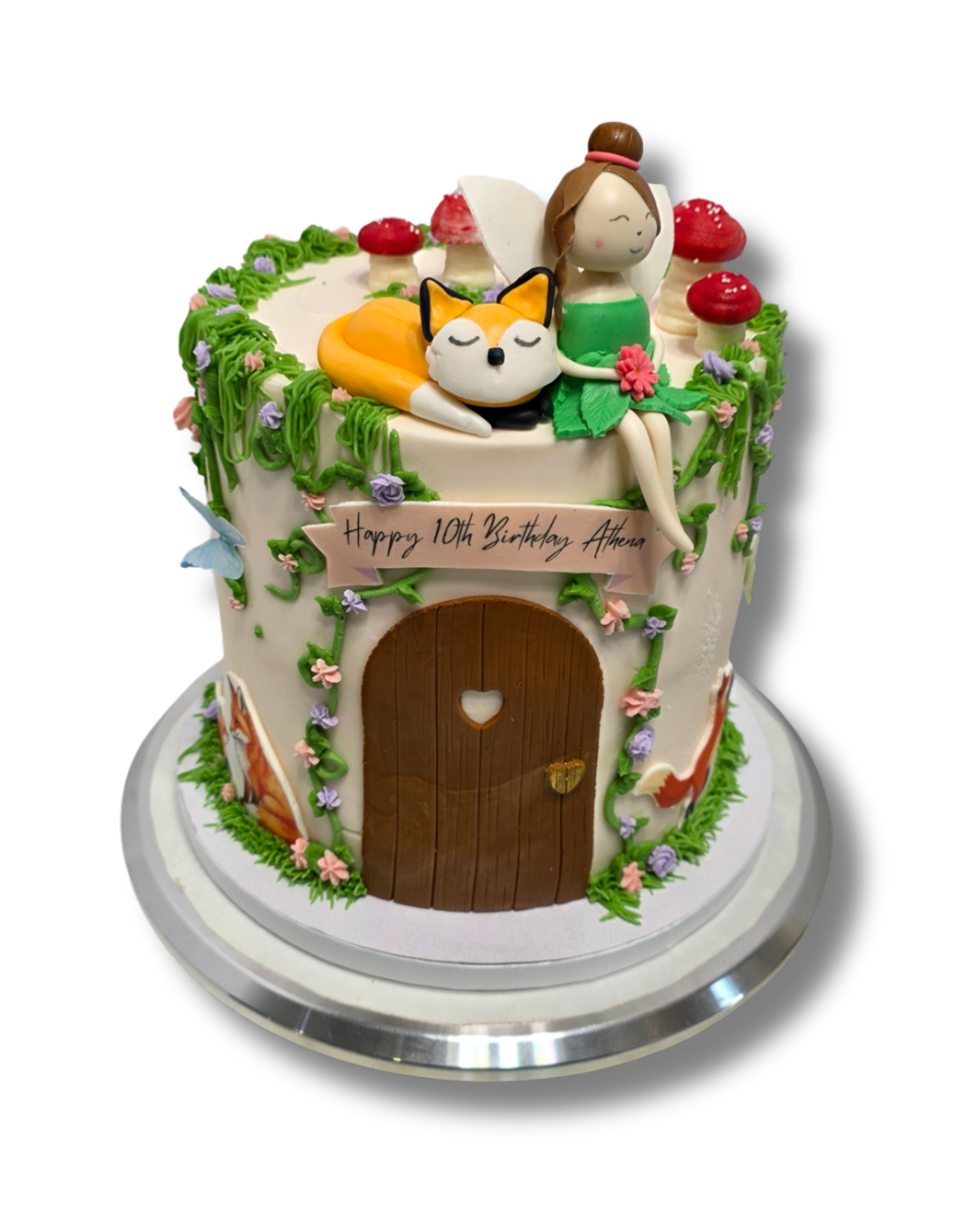 A round fantasy cottage cake decorated with flowers, green vines, and mushrooms, featuring fondant fairy and fox figures, with a door and a sign that reads "Happy 10th Birthday Athena."