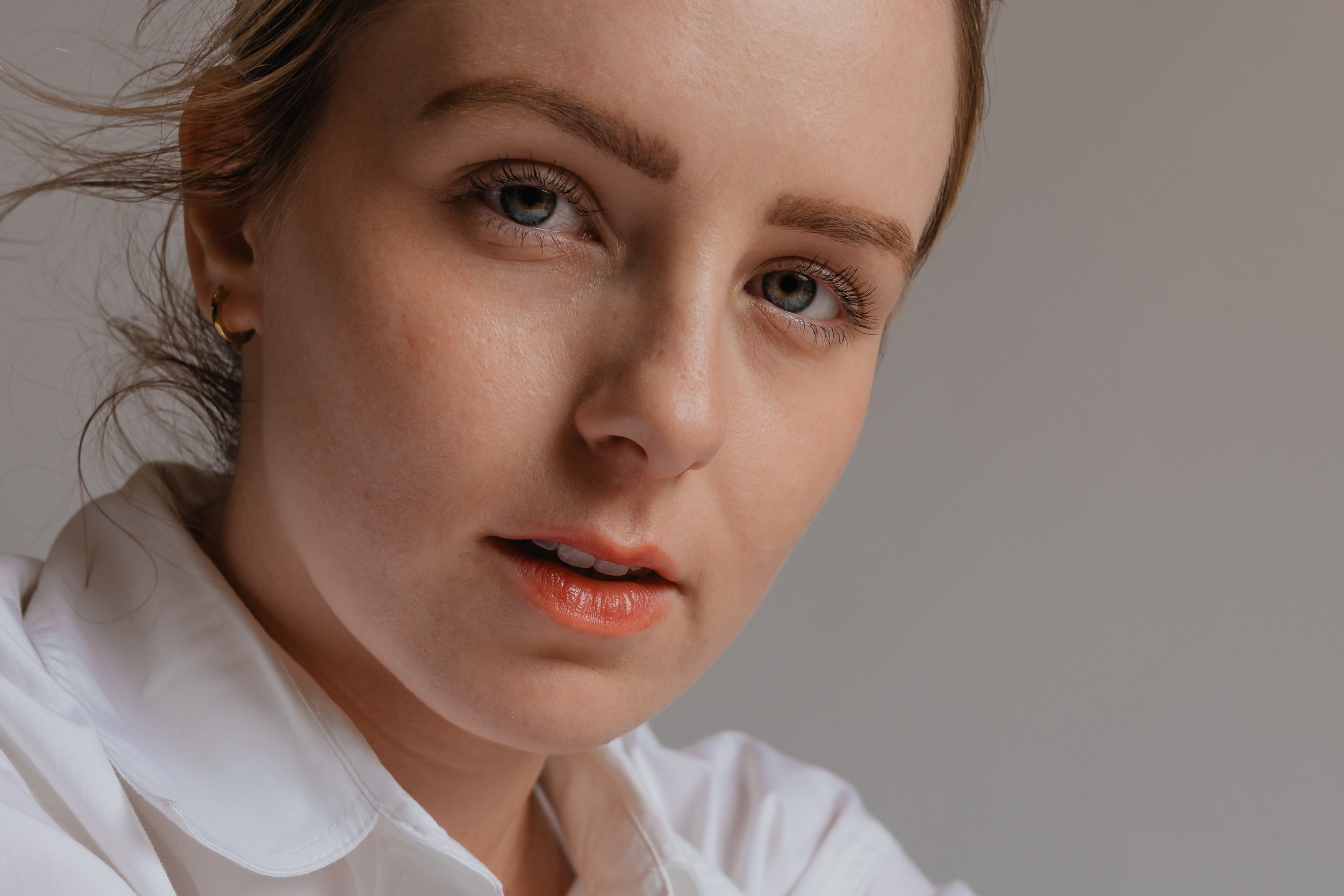 Close-up of a woman with light skin, blue eyes, brown eyebrows, and red hair, wearing a white shirt and small gold hoop earrings, looking at the camera against a neutral background.