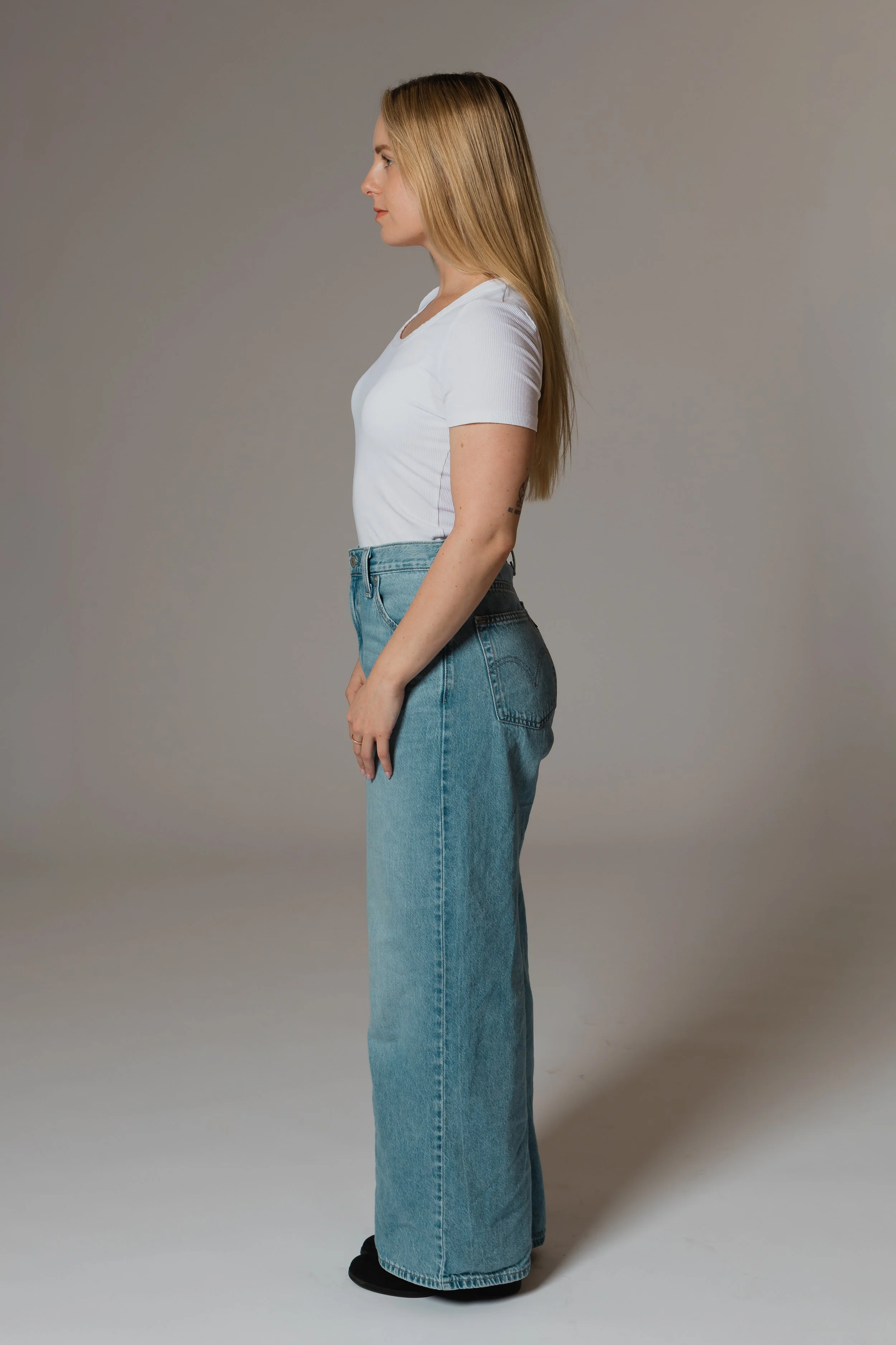 A woman standing in profile against a plain gray background, wearing a white T-shirt, high-waisted blue jeans, and black shoes.