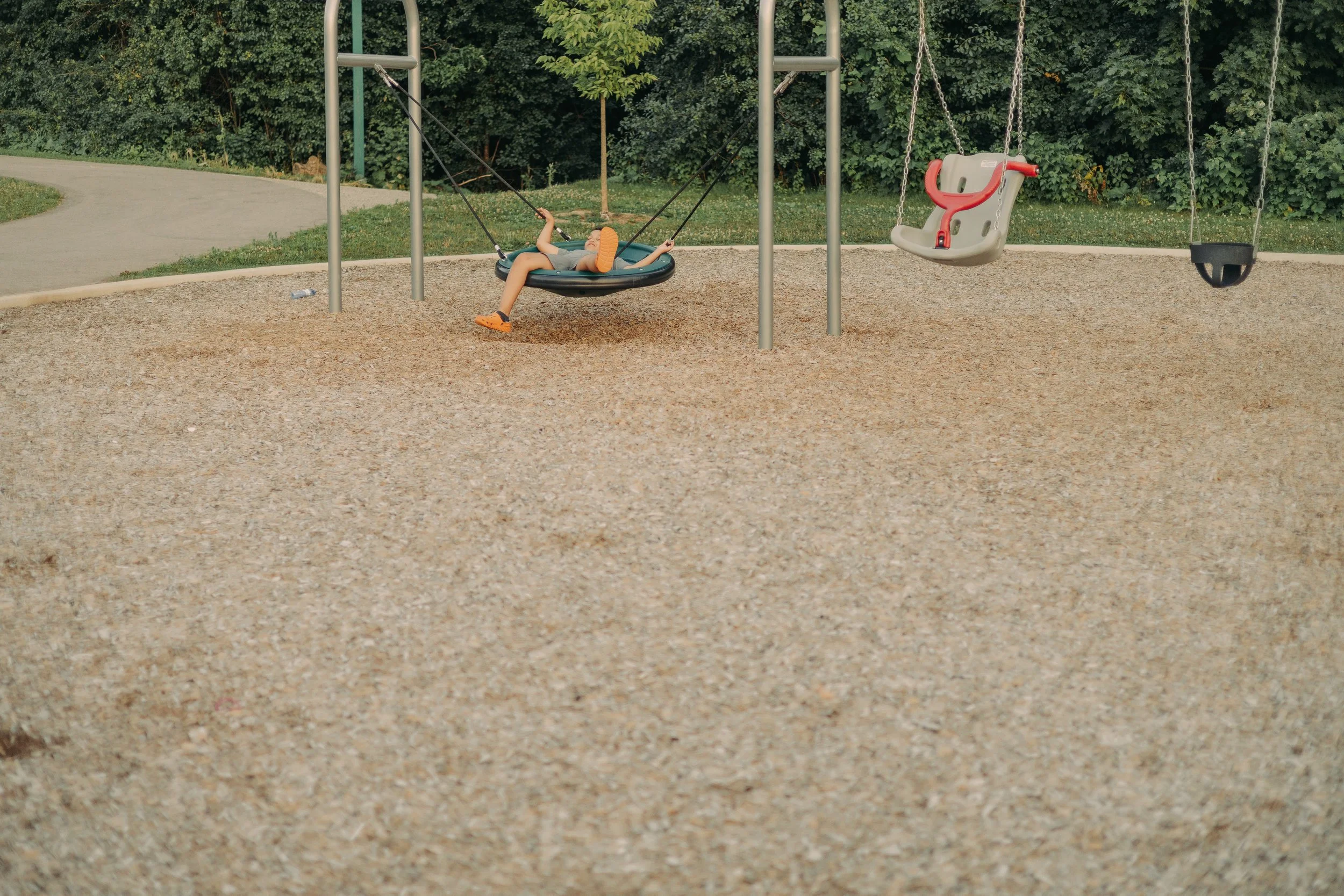 Child lying on a circular toddler swing in a playground with dense green trees in the background.