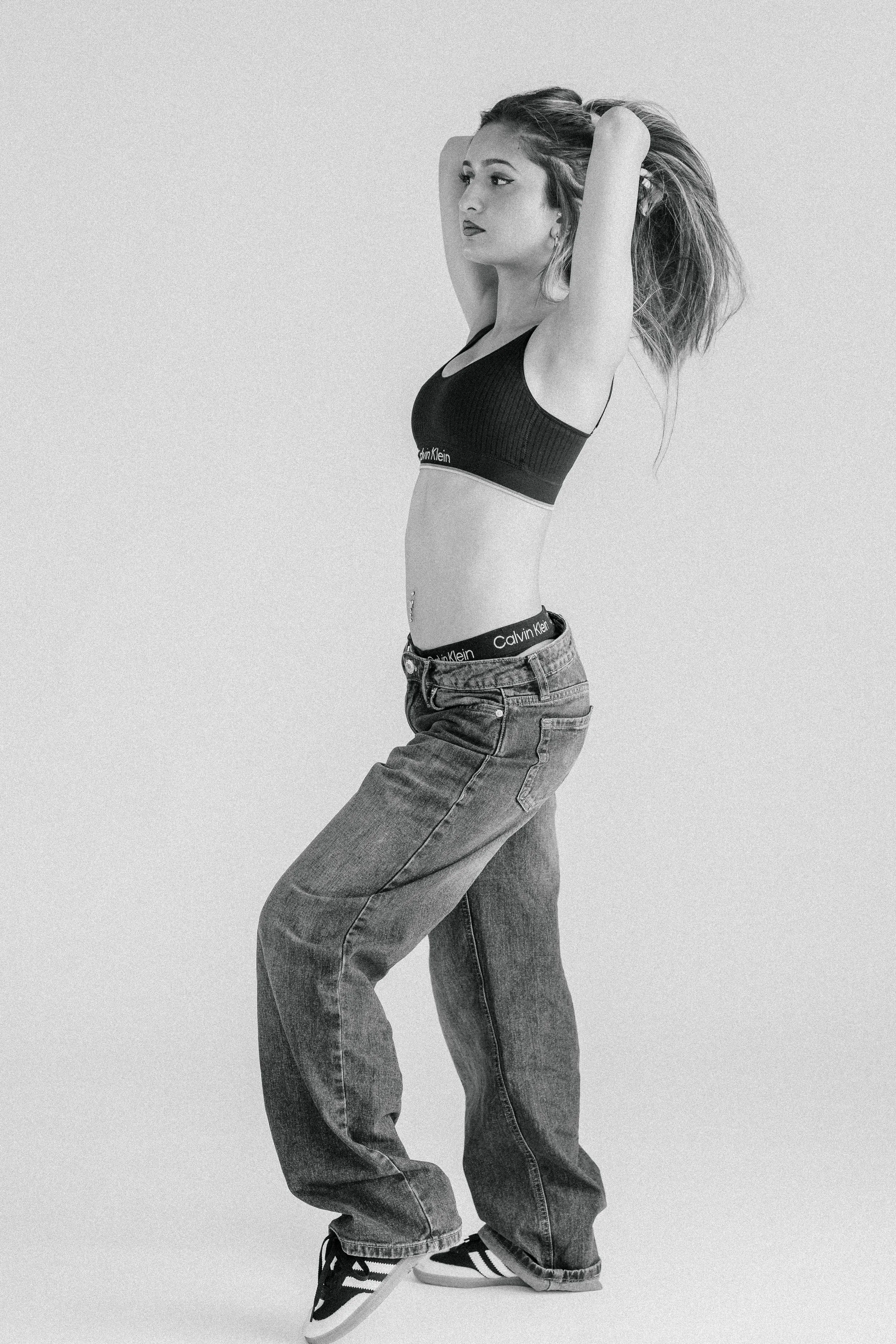 Black and white photo of a young woman with long hair, wearing a sports bra, baggy jeans, and sneakers, standing against a plain background with her arms raised behind her head.