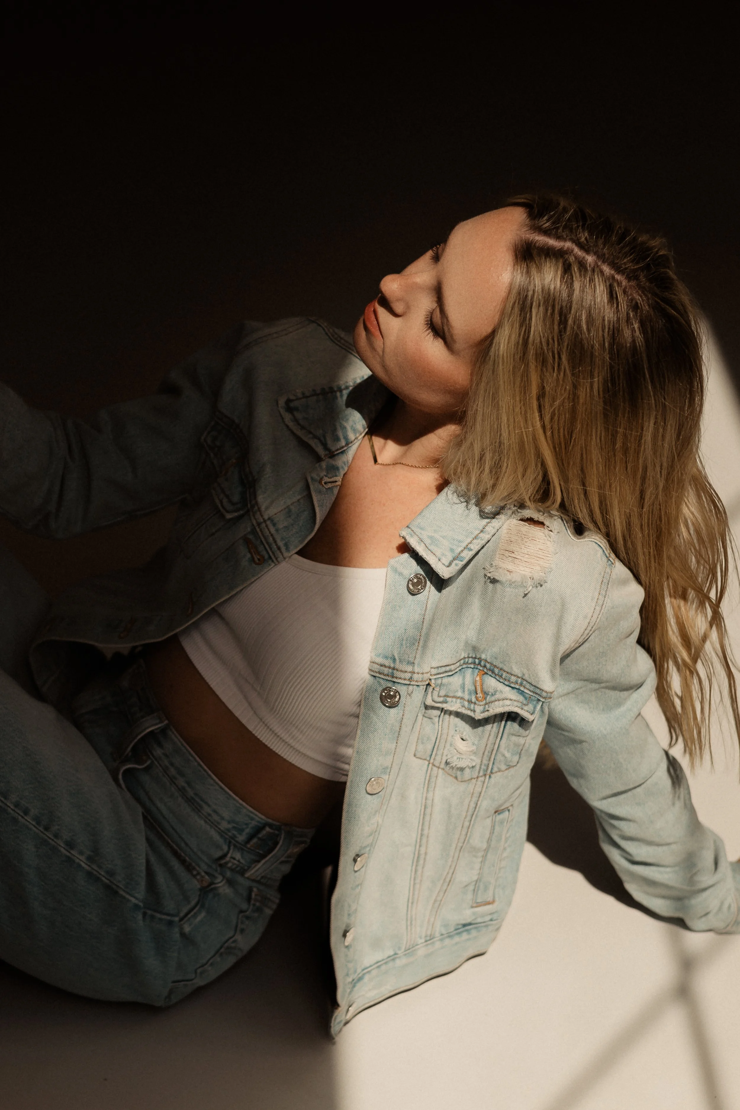 A woman with wavy blonde hair wearing a distressed denim jacket, white crop top, and jeans, sitting against a dark background with her eyes closed.