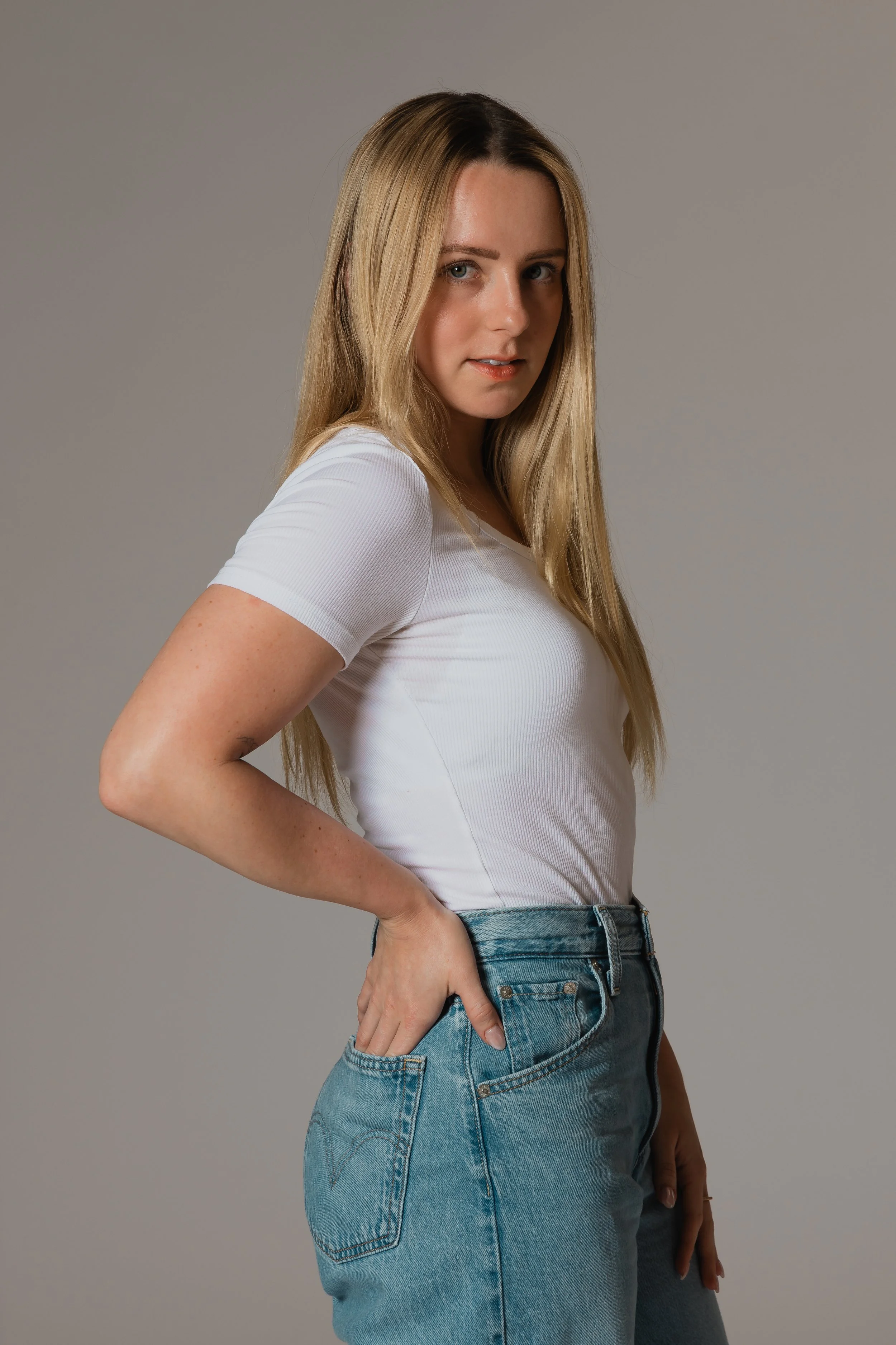 A young woman with long blonde hair, wearing a white t-shirt and blue jeans, standing with her hand on her hip against a plain gray background.