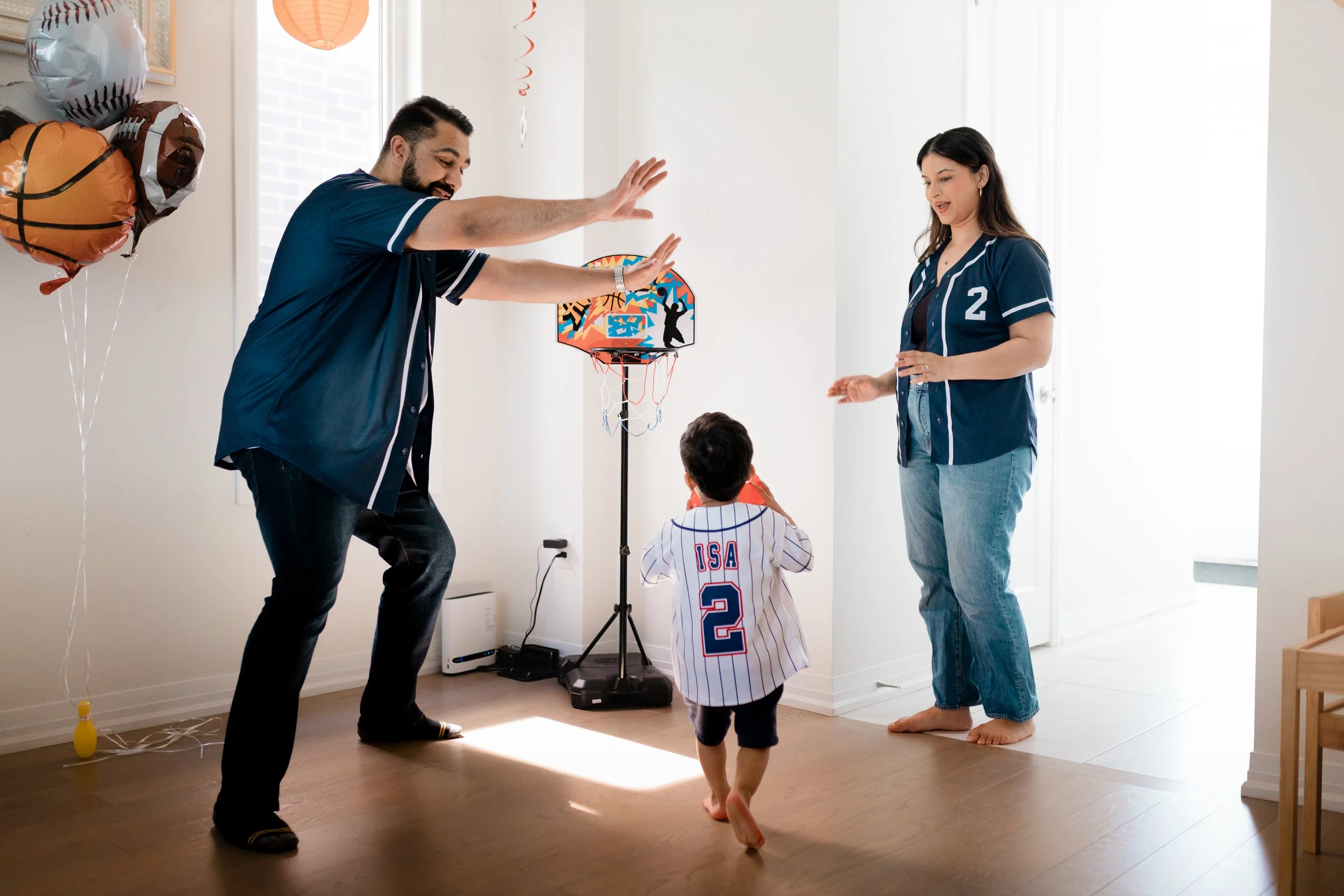 A family celebrating a birthday indoors with balloons and a small basketball hoop. A man and woman are playing with two children, one in a baseball jersey. The man is giving a high five to the child in the jersey, while the woman and the other child watch and smile.