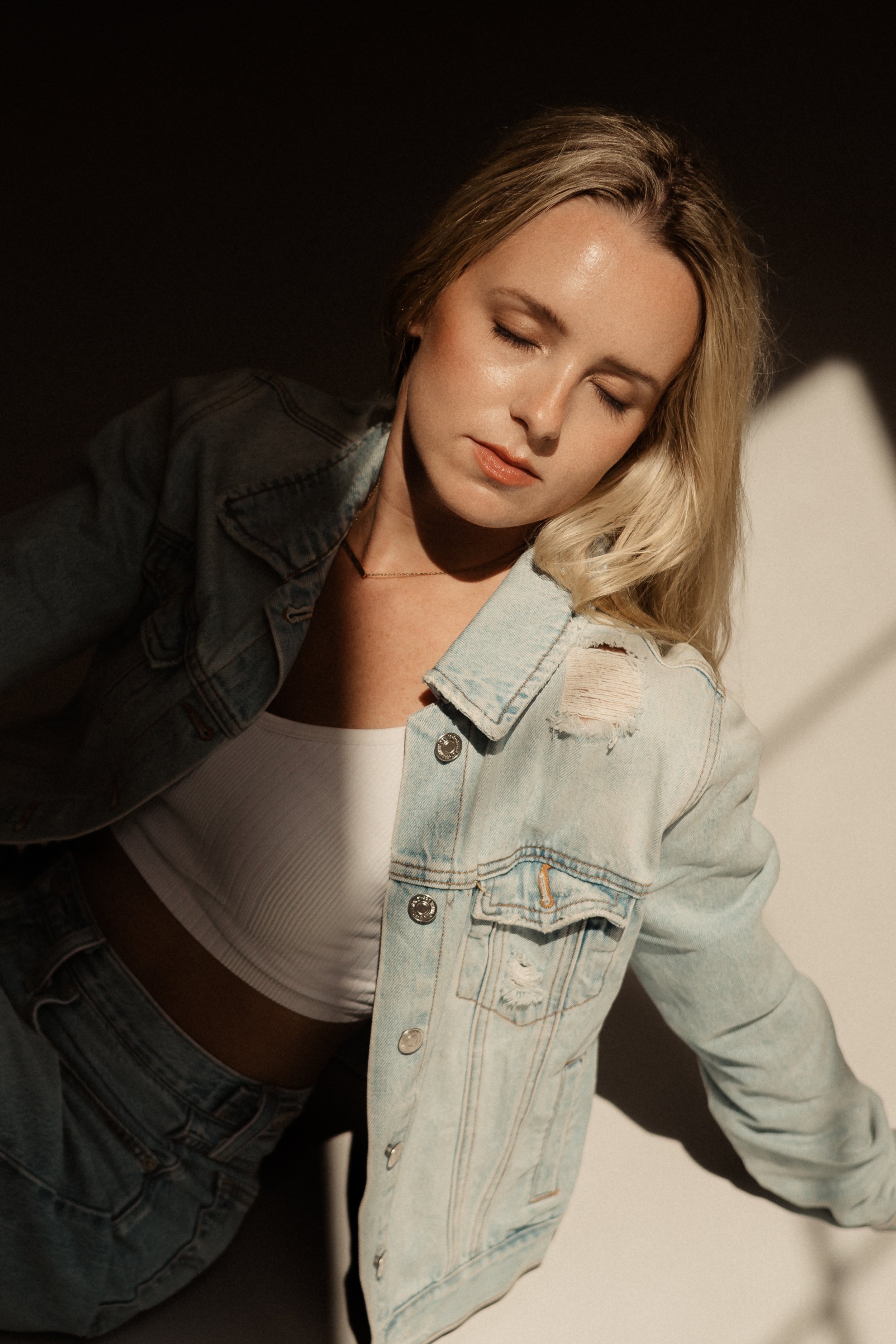 A woman with blonde hair and closed eyes wearing a denim jacket over a white crop top, standing against a dark background.