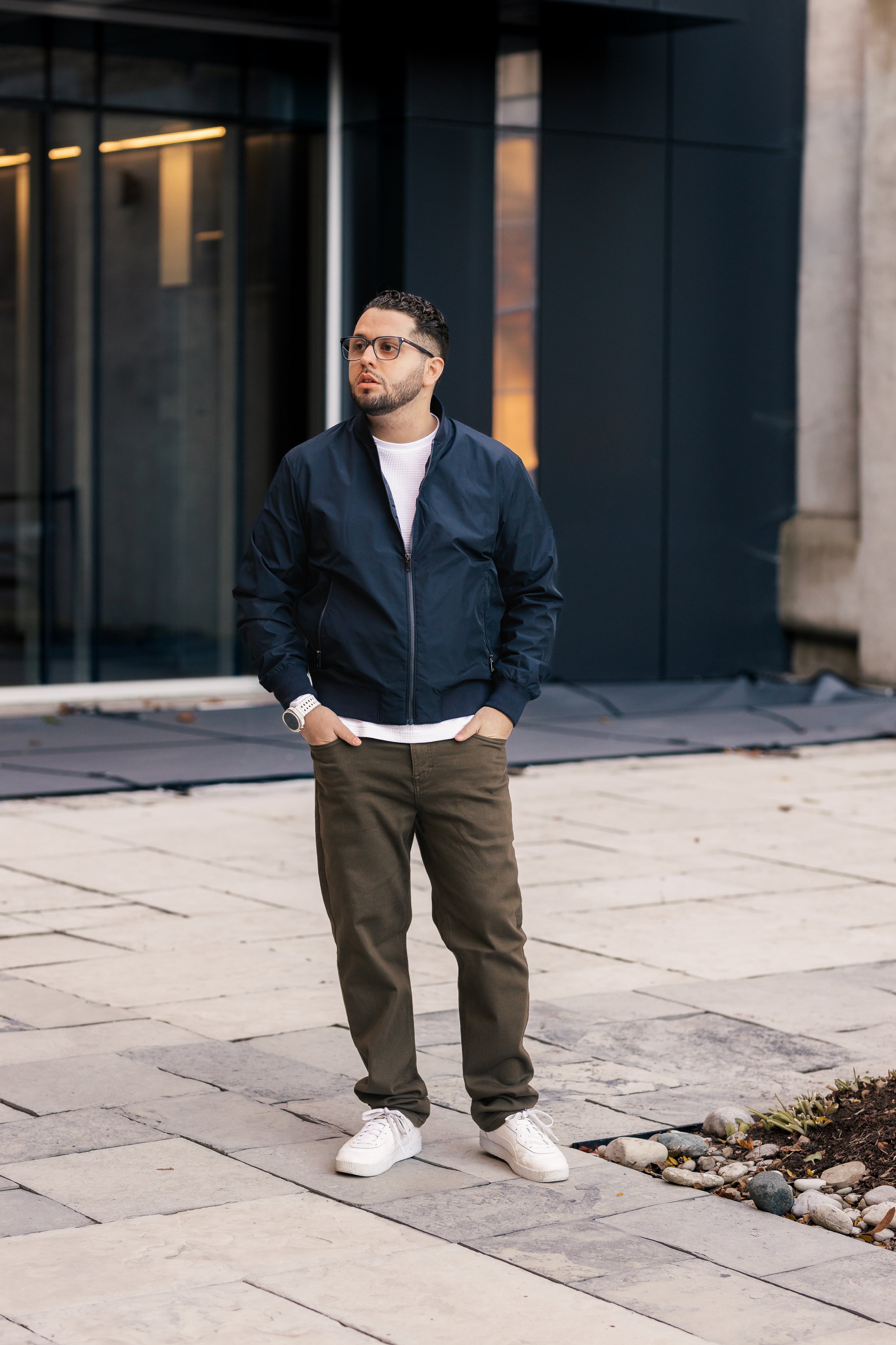 A young man and woman standing outdoors in snowy landscape, wearing matching green Christmas sweaters with reindeer and trees, black beanies with reindeer antlers, and winter boots, smiling at the camera.